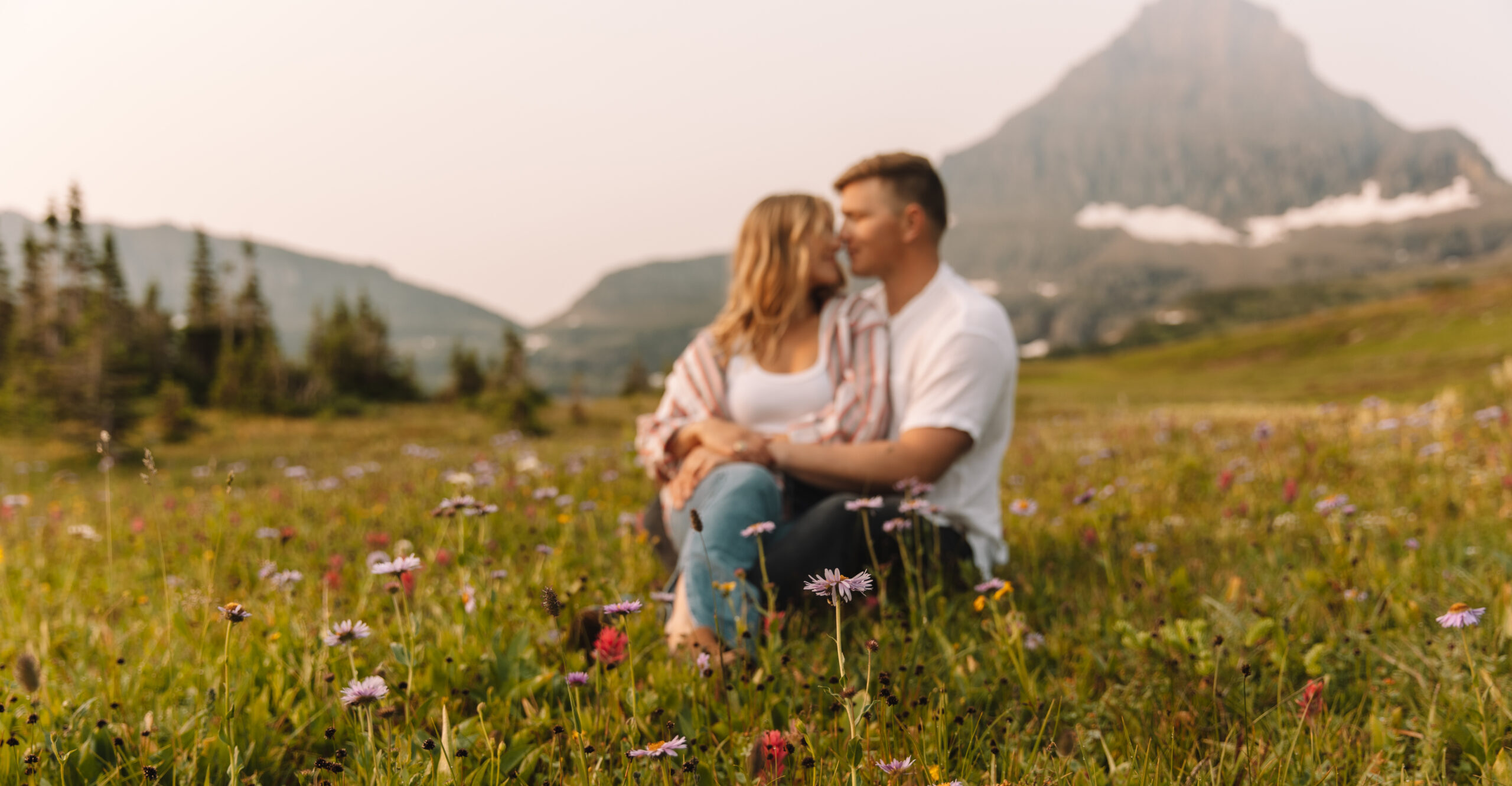Girl sitting on guys lap between a field of wild flowers with the mountains in the background in Glacier national Park
