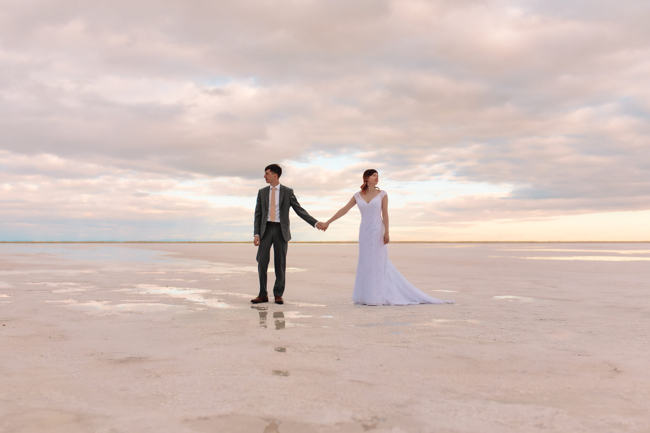 Elopement couple standing next to each other holding hands, looking in opposite directions on the flooded salt flats in Utah. The candy floss skies are reflecting in the water below them