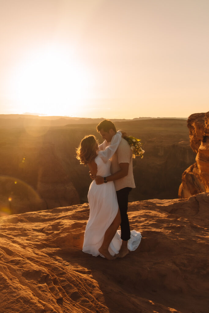 Groom holding his bride around her waist while her arms are around his neck as the sun sets at Horse Shoe Bend in Arizona