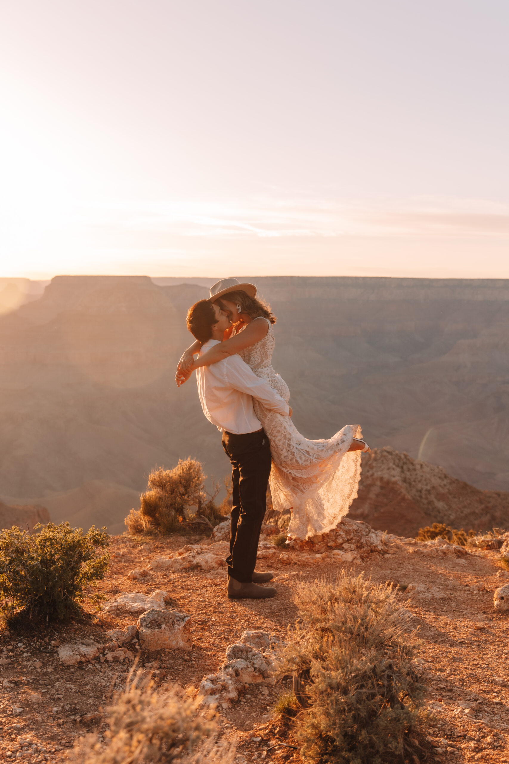 Groom picking up his bride standing on the edge of a cliff at The Grand Canyon in Arizona with the vast canyons in the background. Her leg lifts up passionately as they kiss. She is wearing a hat and a lace wedding dress