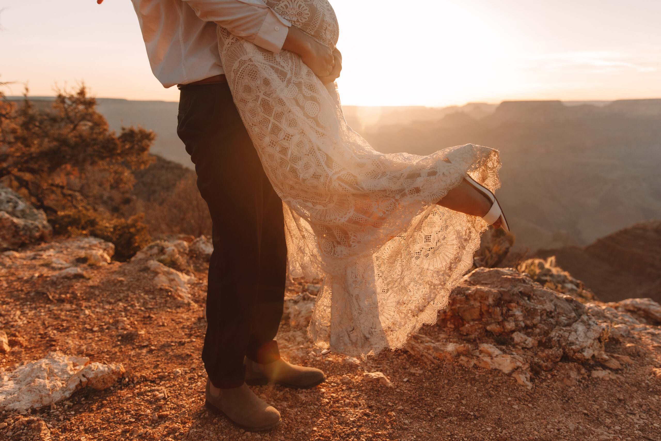 A close up shot of the bottom halves of a groom picking up his bride as she kicks her leg back. The sun shines through her lace wedding dress and her white high heel shoes are visible