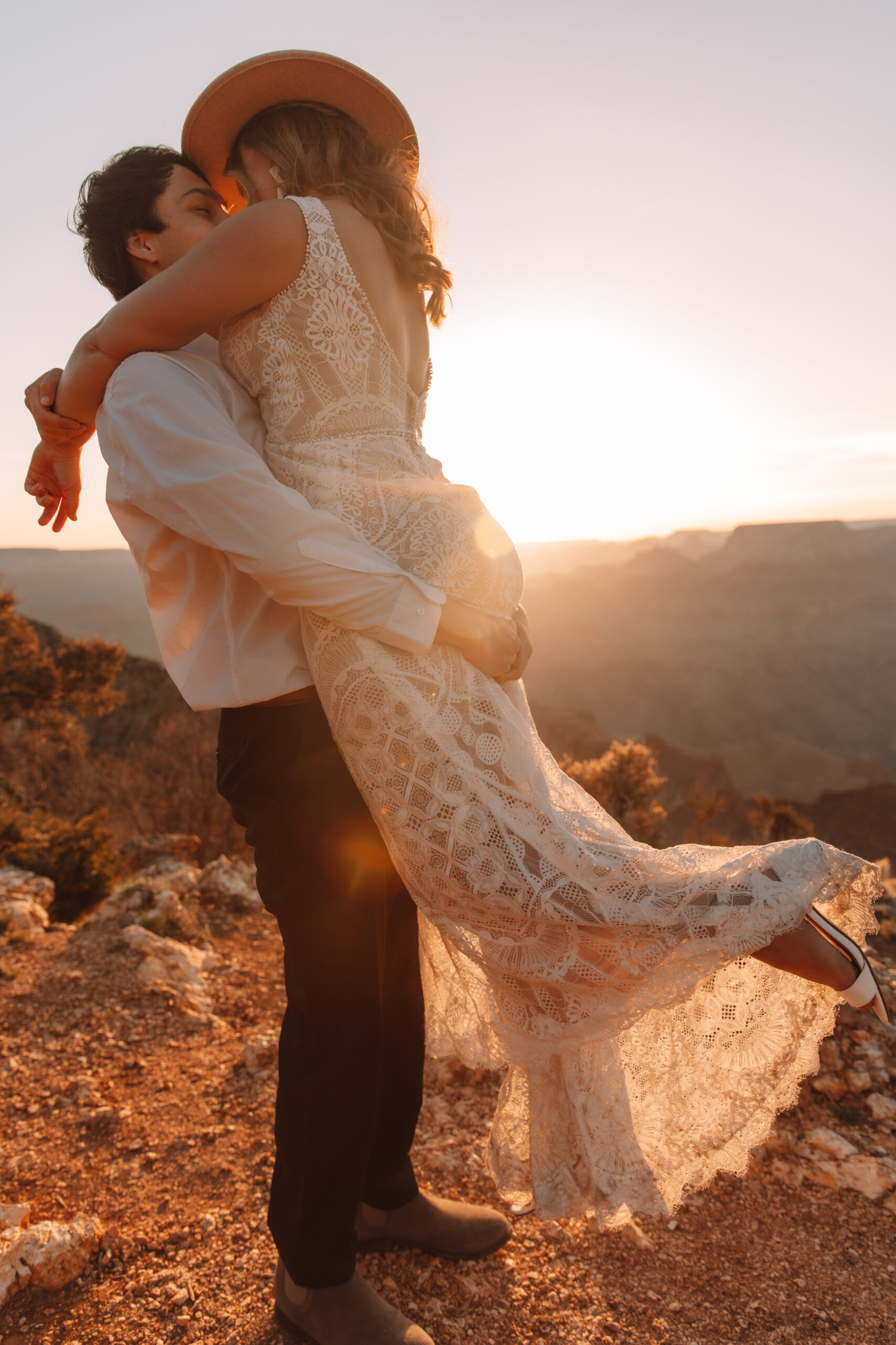Groom picking up his bride standing on the edge of a cliff at The Grand Canyon in Arizona with the vast canyons in the background. Her leg lifts up passionately as they kiss. She is wearing a hat and an open back  lace wedding dress