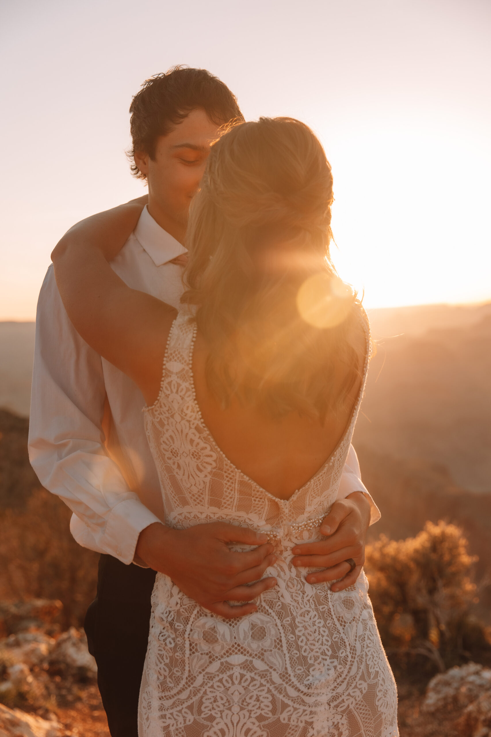 A groom holding his bride while her back is visible to us in her stunning lace open back wedding dress. The sun is setting behind them in the Grand Canyon in Arizona