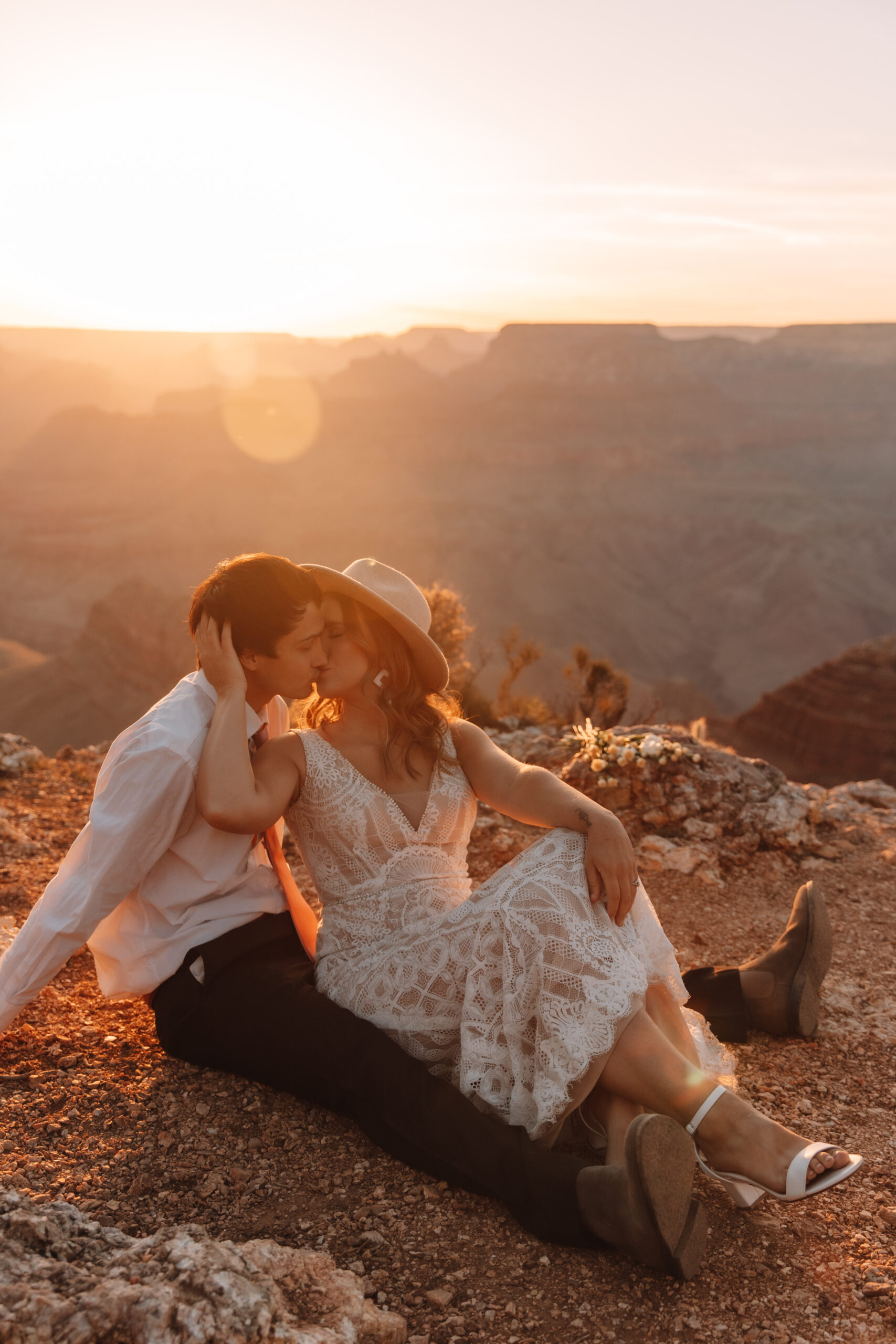 A bride and groom sitting on a cliff in the Grand Canyon as the sun sets behind them. The bride is cupping her groom's face as they kiss. She is wearing a hat and a lace dress. He is wearing a white button up long sleeve shirt with a pink tie
