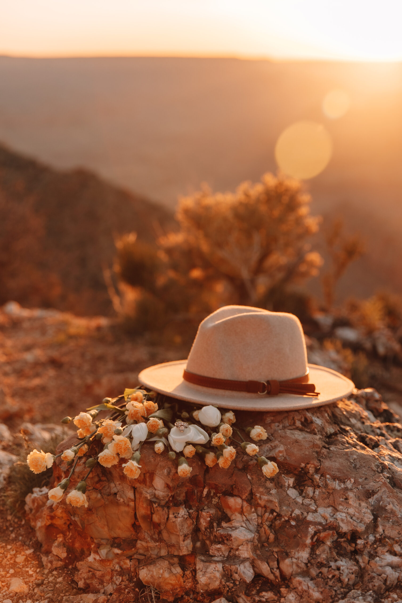 Stunning detail photo of a hat lying next to a bouquet of yellow and white flowers on top of a rock in the Grand Canyon as the sun sets behind the canyons. The bride's wedding ring is lying on top of one of the flowers
