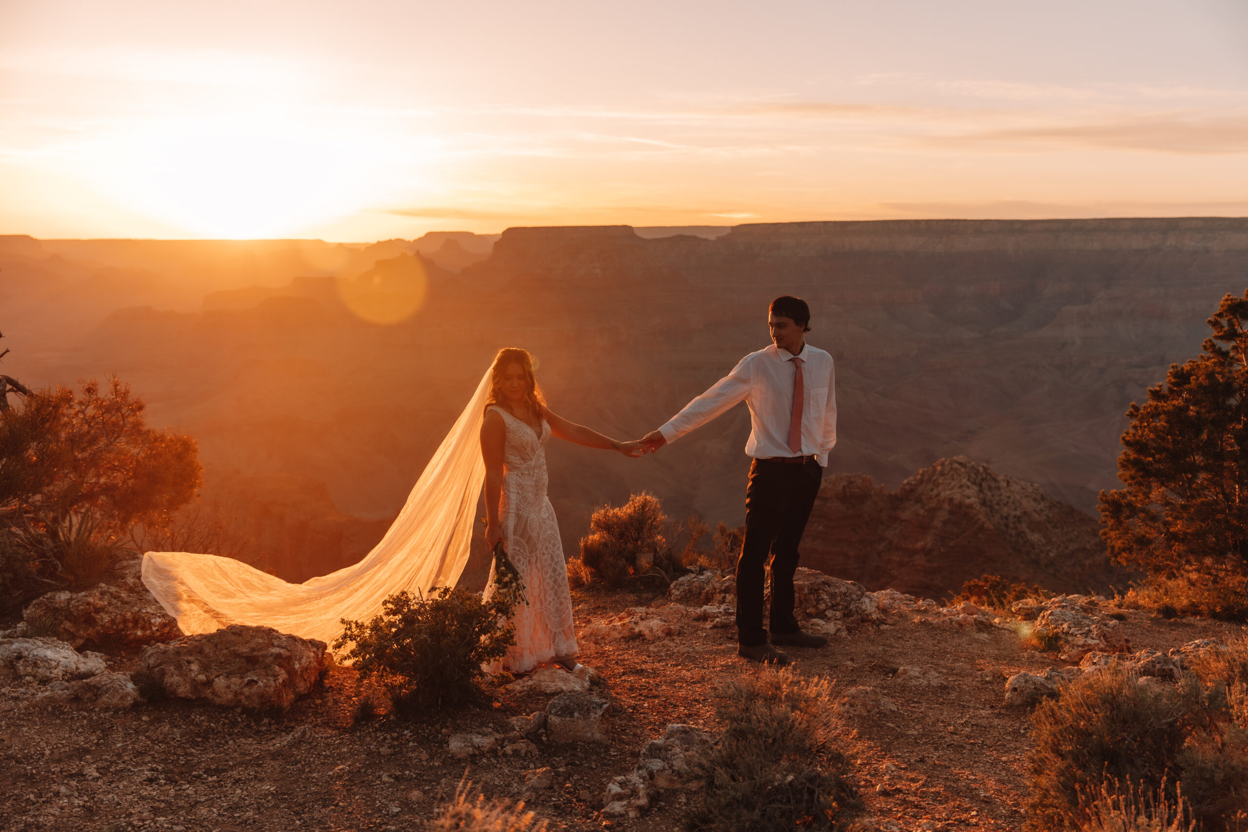 A groom walking in front of his bride, while he's holding her hand and leading her across the cliff as the sun sets behind them. Her veil is long and flowing behind her. She is wearing a beautiful lace elopement dress