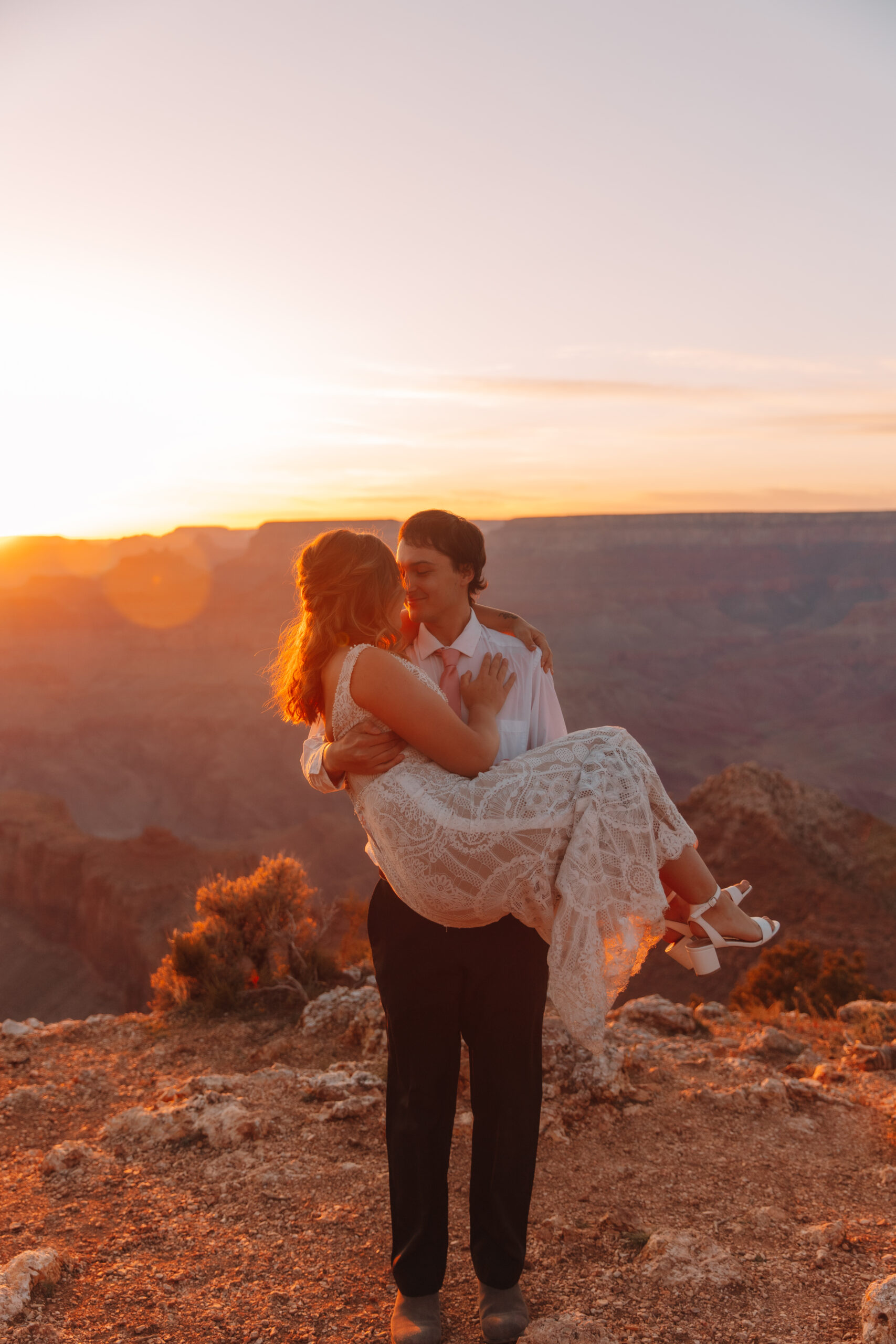 A groom picking up his bride and holding her like a baby as they walk across the cliff in the Grand Canyon. The sun is setting behind them