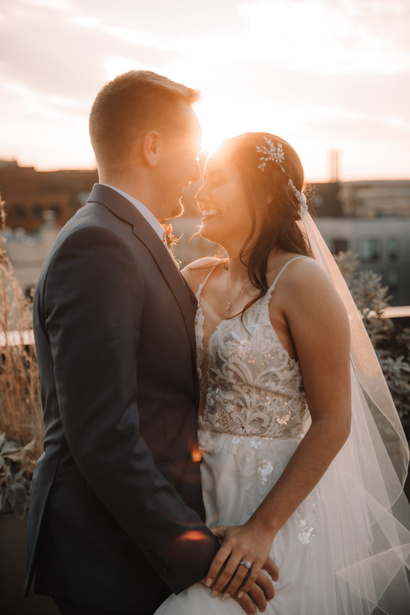 Bride and groom facing each other laughing on top of a deck overlooking the city as the sun sets behind them