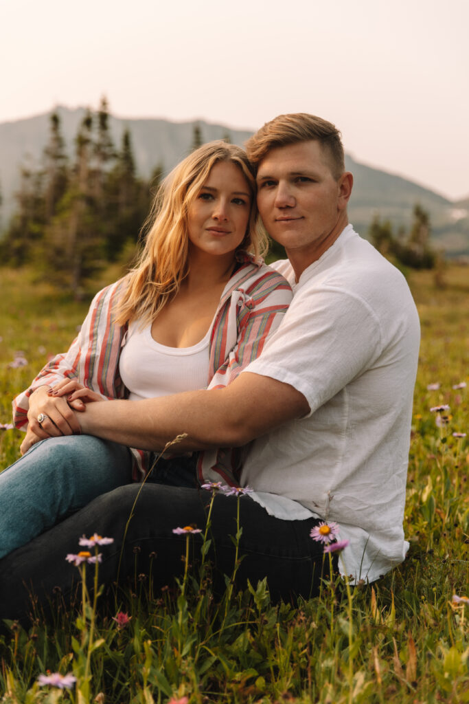 Newly engaged couple sitting in between the grass with wild flowers around them staring into the camera 