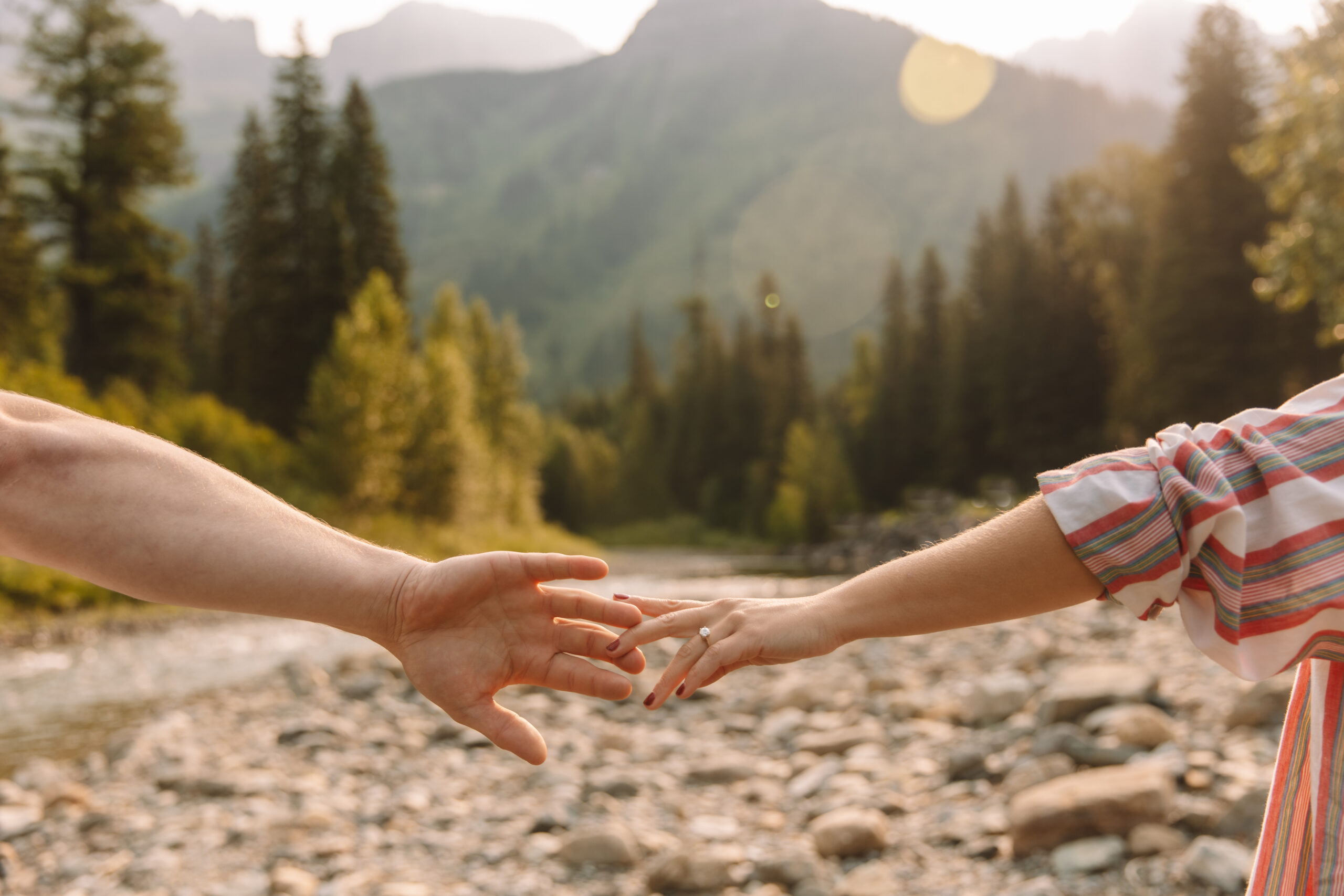 Newly engaged couple reaching out their hands to each other with the mountains in the background