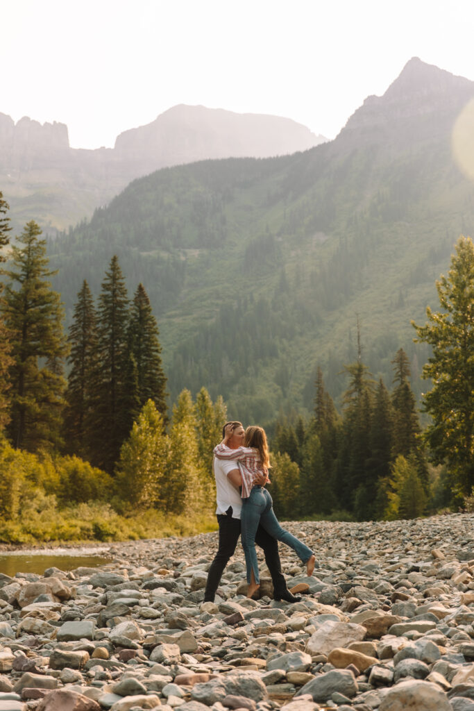 Newly engaged couple kissing in Glacier National park, down by the river with the mountains in the back
