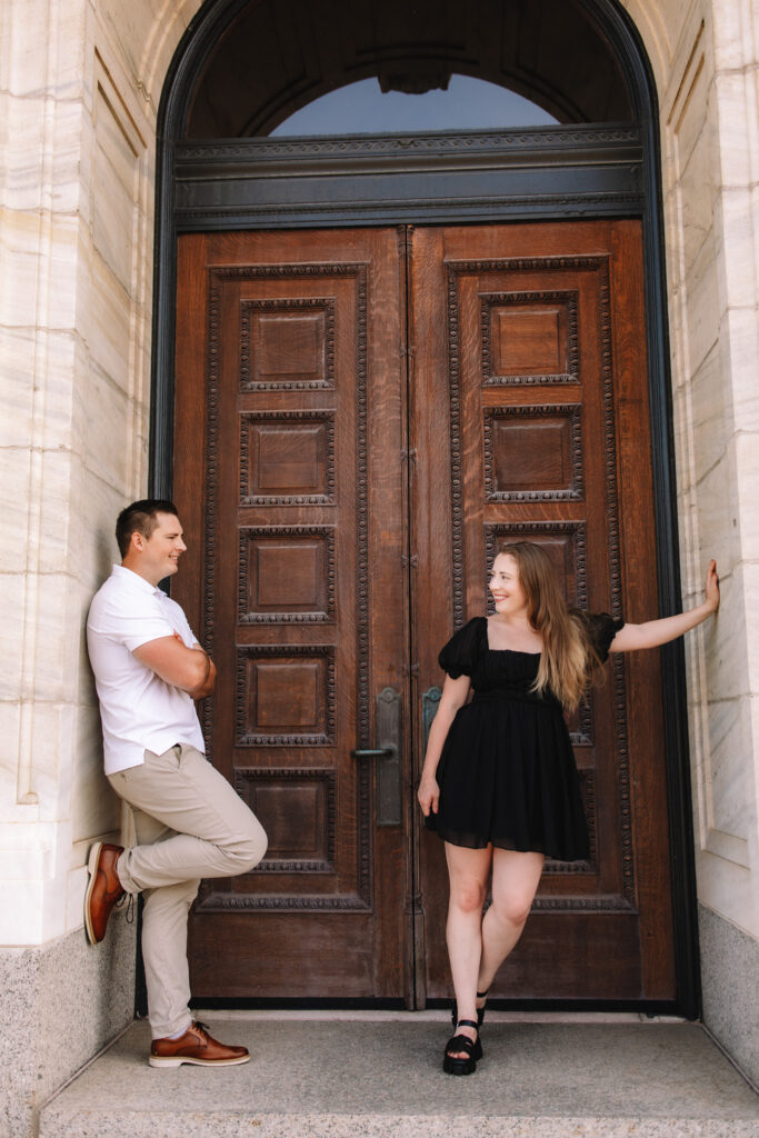 Guy leaning against the wall in front of a large wooden door, admiring his beautiful lady who is dressed in a black dress also leaning against the wall opposite from him