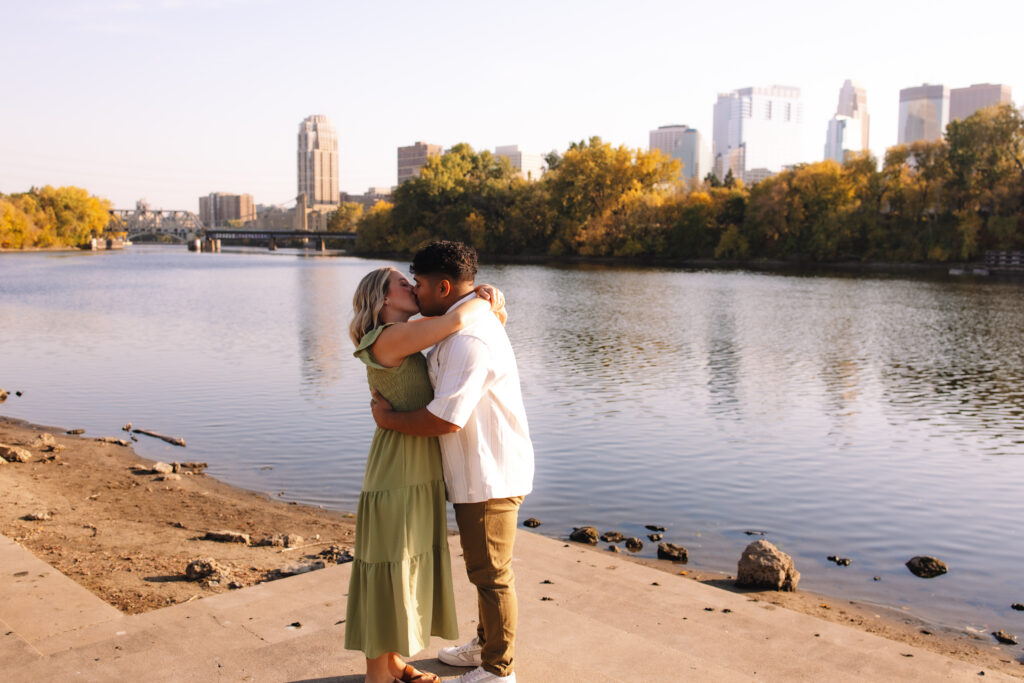 Girl wrapping her arms around her fiancé's neck as they kiss tenderly with the river and Minneapolis skyline in the background