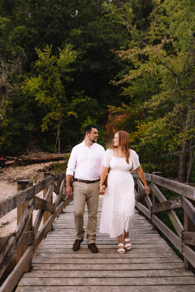 Engagement couple dressed in formal attire walking across a wooden bridge at Minihaha Falls in Minneapolis