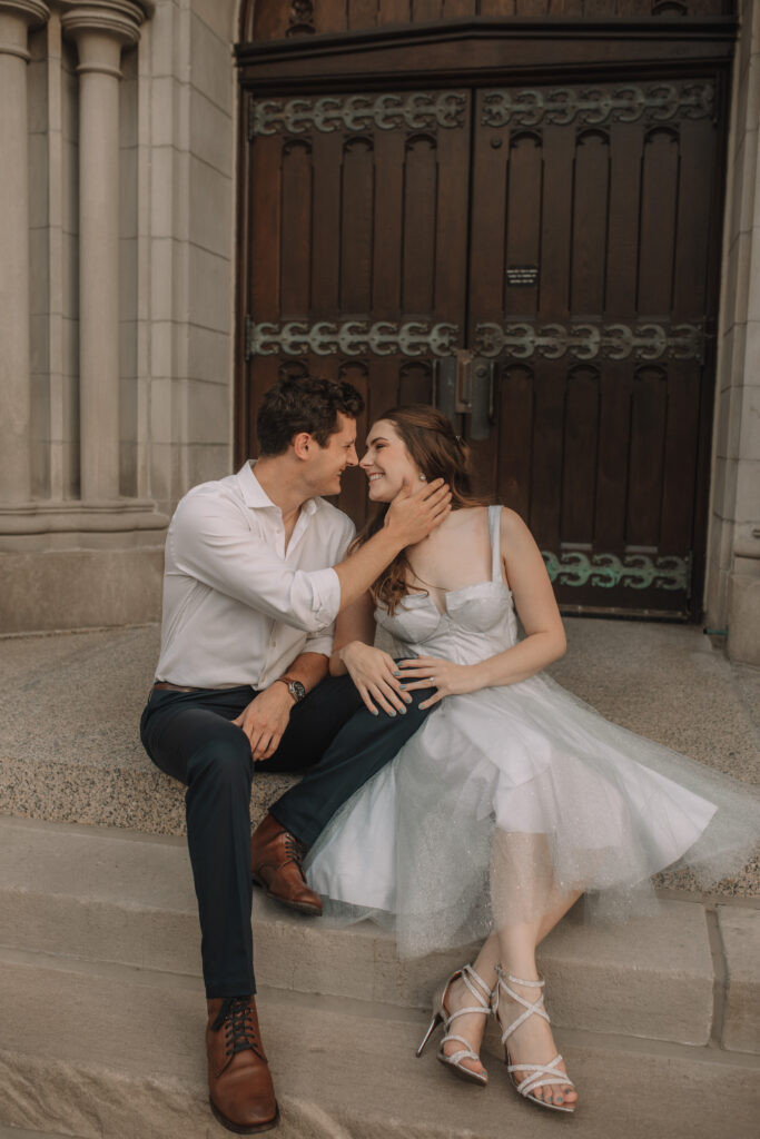 An engagement couple in formal attire sitting on the stairs of the American Institute of Art building while the guys gently grabs his fiancés face to pull her in for a kiss