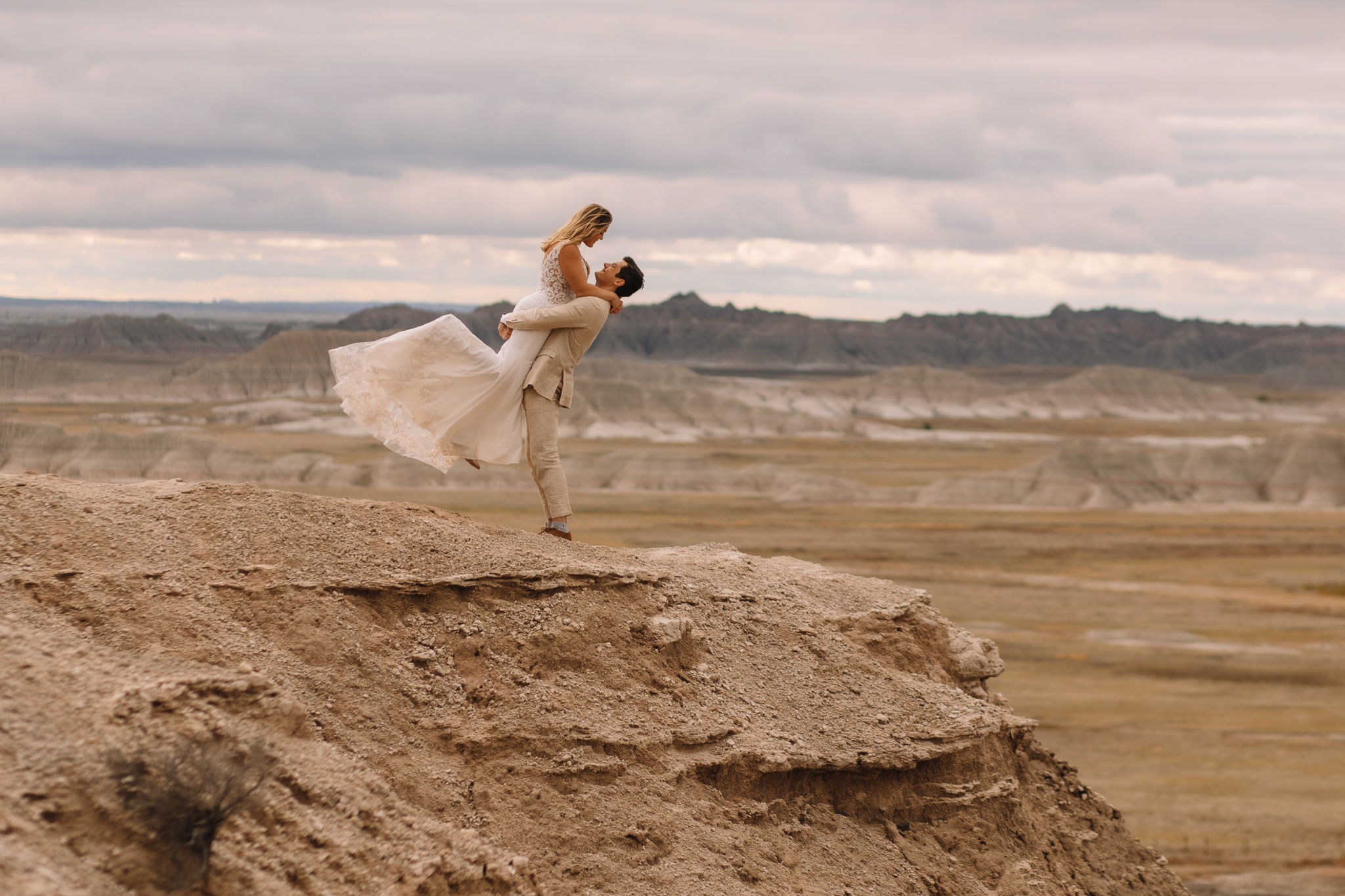 Groom picking up his bride from below her butt as she kicks her legs up and down and her dress is flowing in the wind. The South Dakota badlands are visible around them