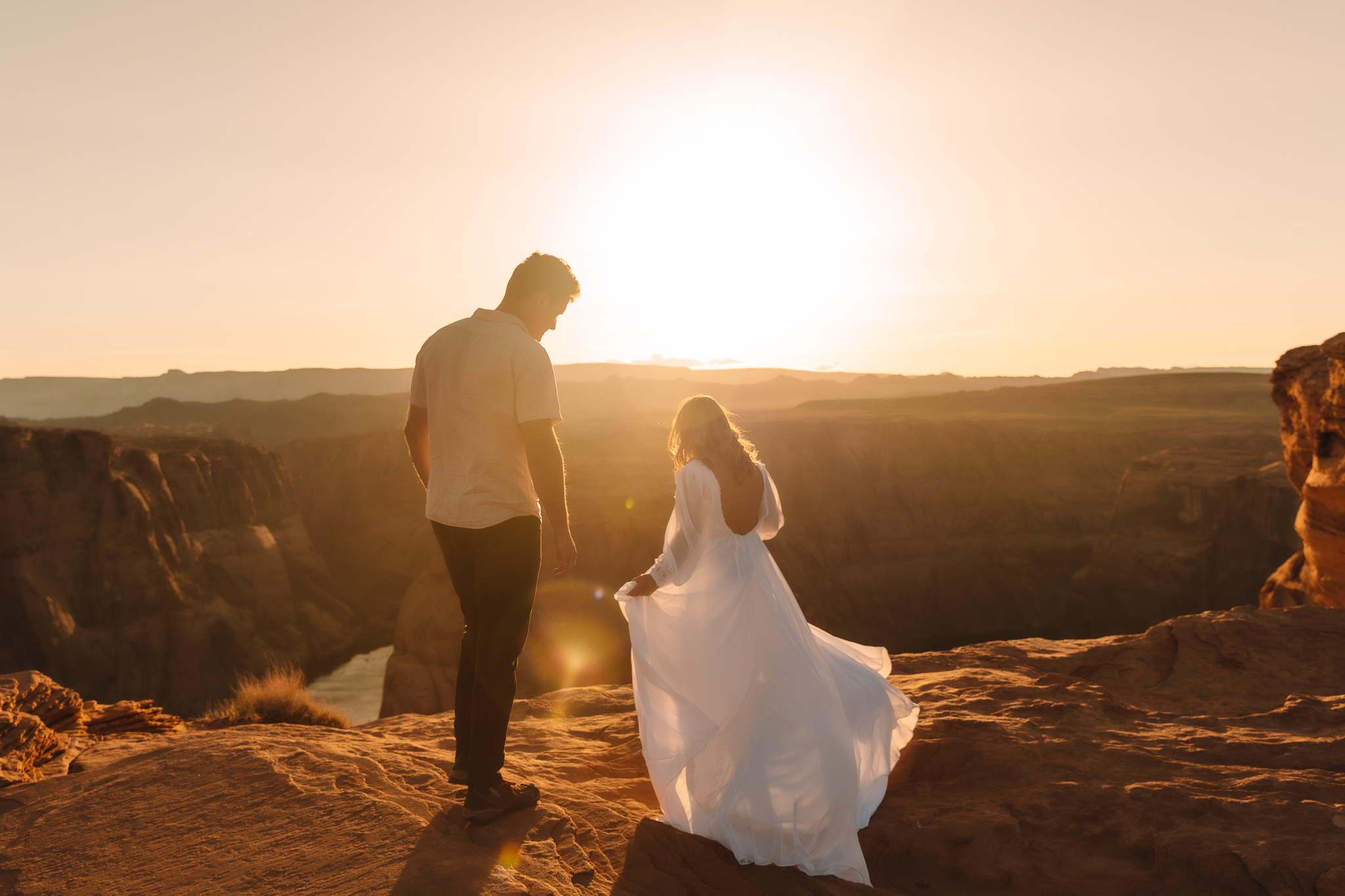A bride in a Flowing, chiffon hiking elopement dress standing on the edge of a cliff with her groom at Horseshoe bend in Arizona