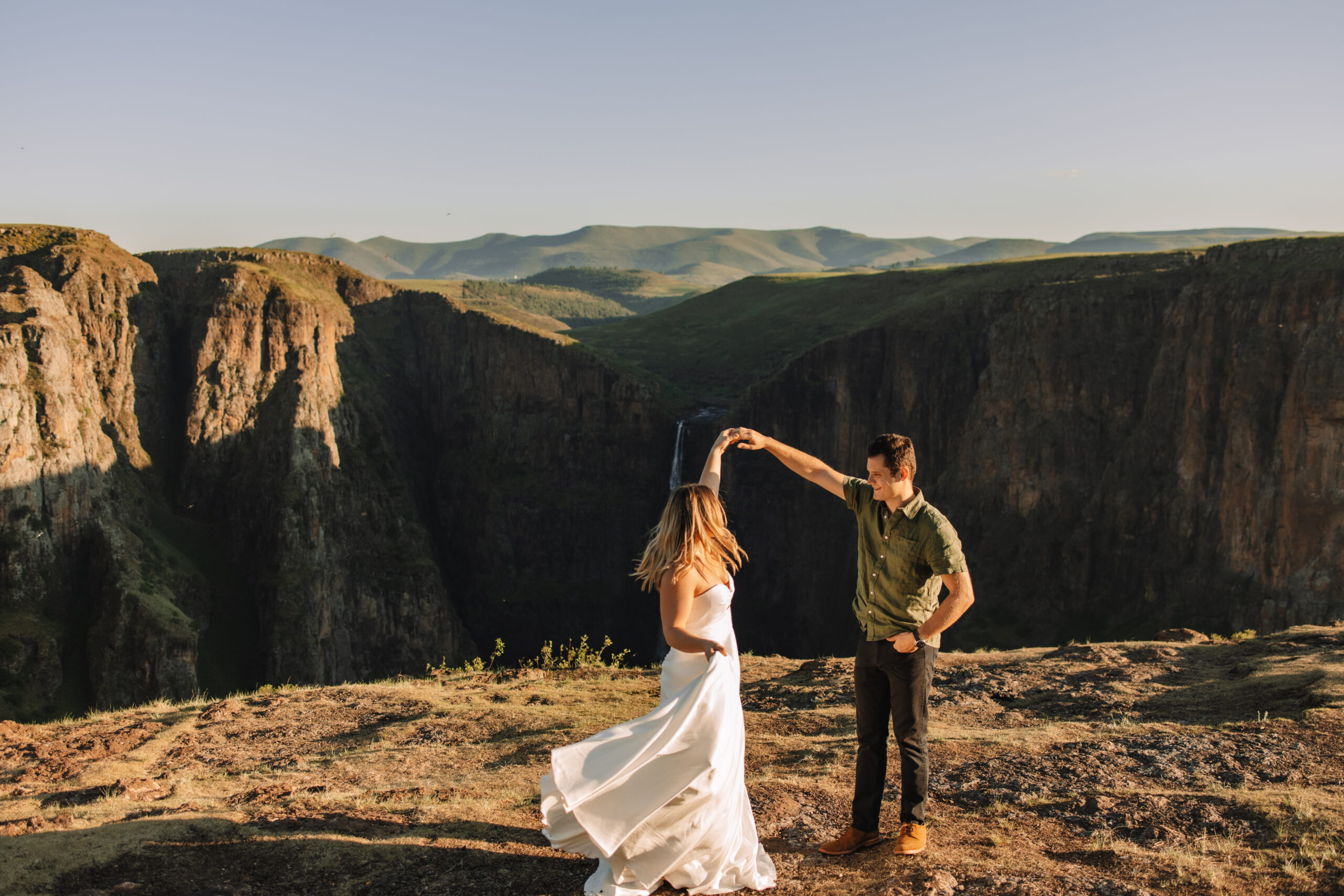 South Africa elopement couple standing on top of a cliff in the Drakensberg while groom twirls his bride. A beautiful waterfall is visible in the back ground