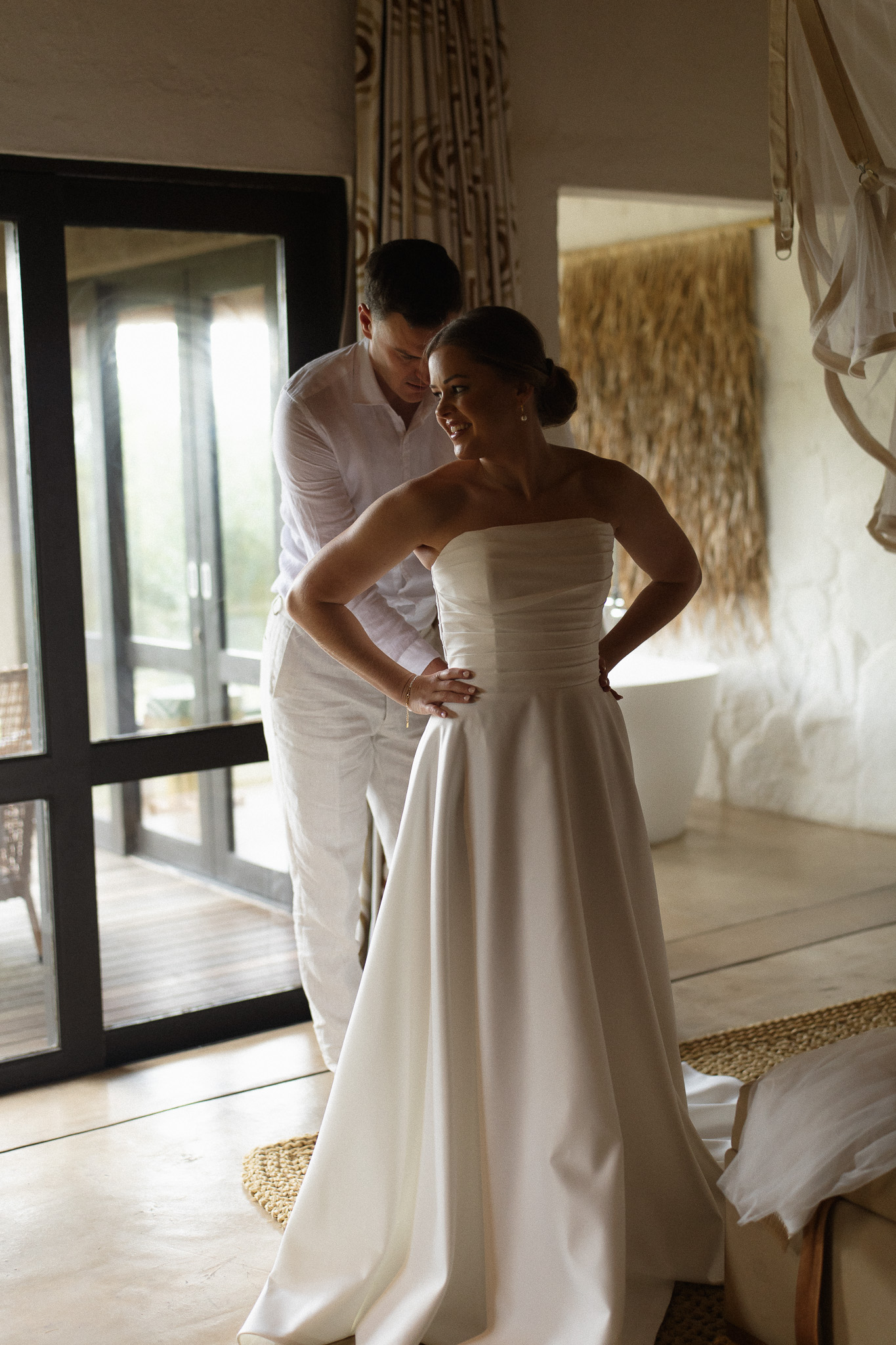Groom helping the bride fasten her wedding dress inside a safari lodge suite