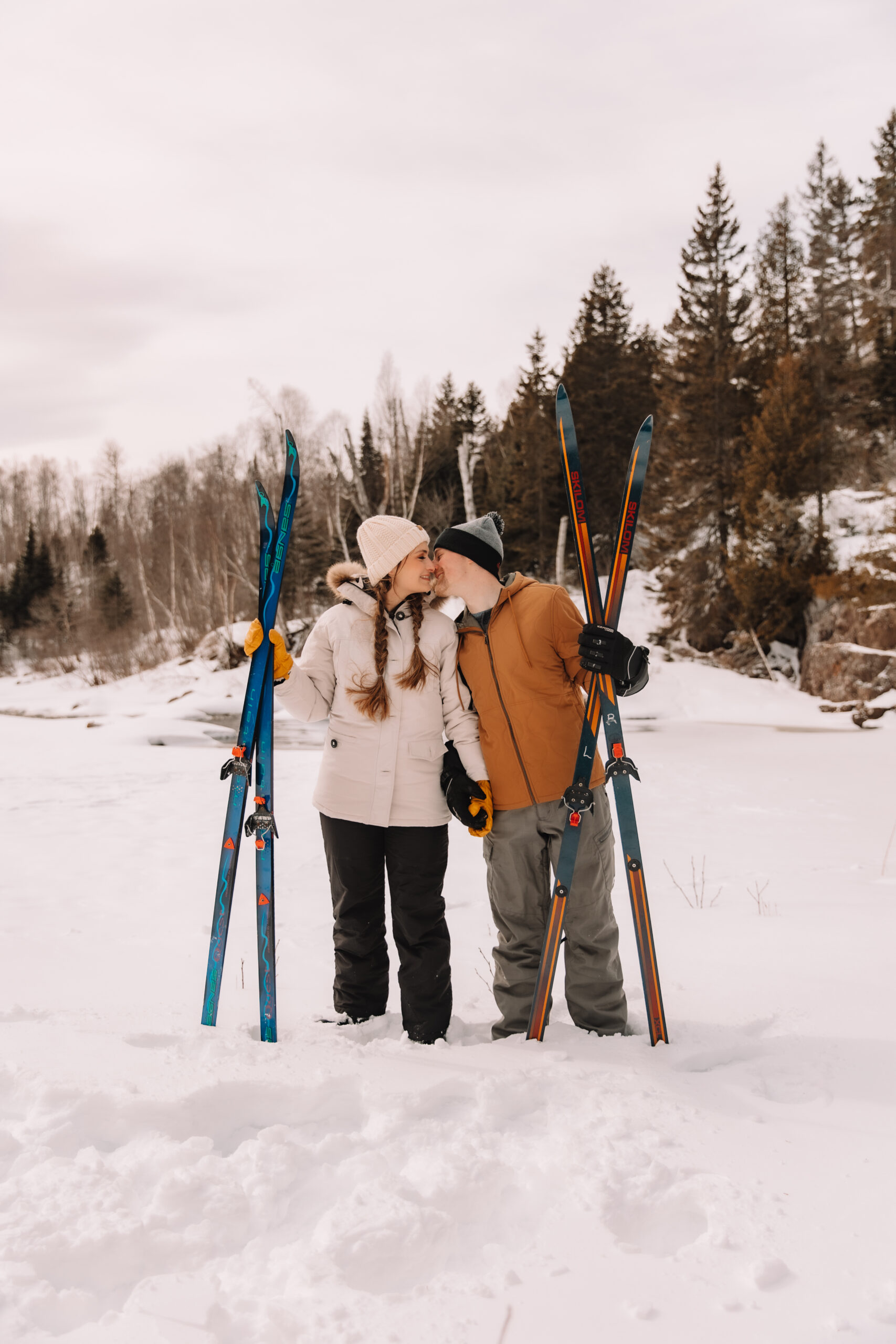 A couple dressed up in warm winter attire as they stand next to each other in the snow with their skis next to them planted in the snow