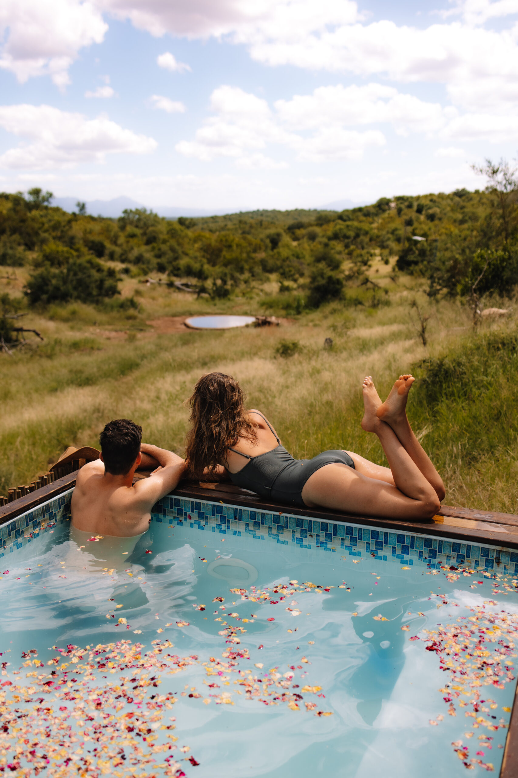 A honeymoon couple relaxing by a pool with flowers floating around them as they overlook the trees 