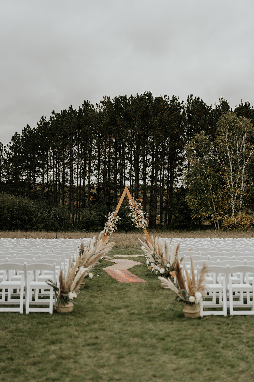 Outdoor wedding setup with white chairs and a triangular wooden arch, surrounded by lush trees. decorative grasses flank a central aisle.