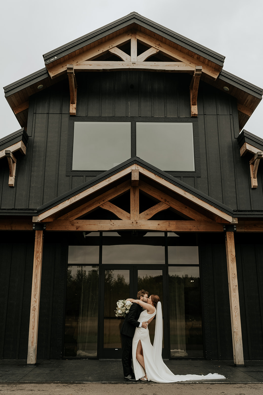 Exterior view of a modern wedding venue black wooden barn-style house with large windows, surrounded by a grassy lawn and overcast sky. Thus venue is called Ivy Black
