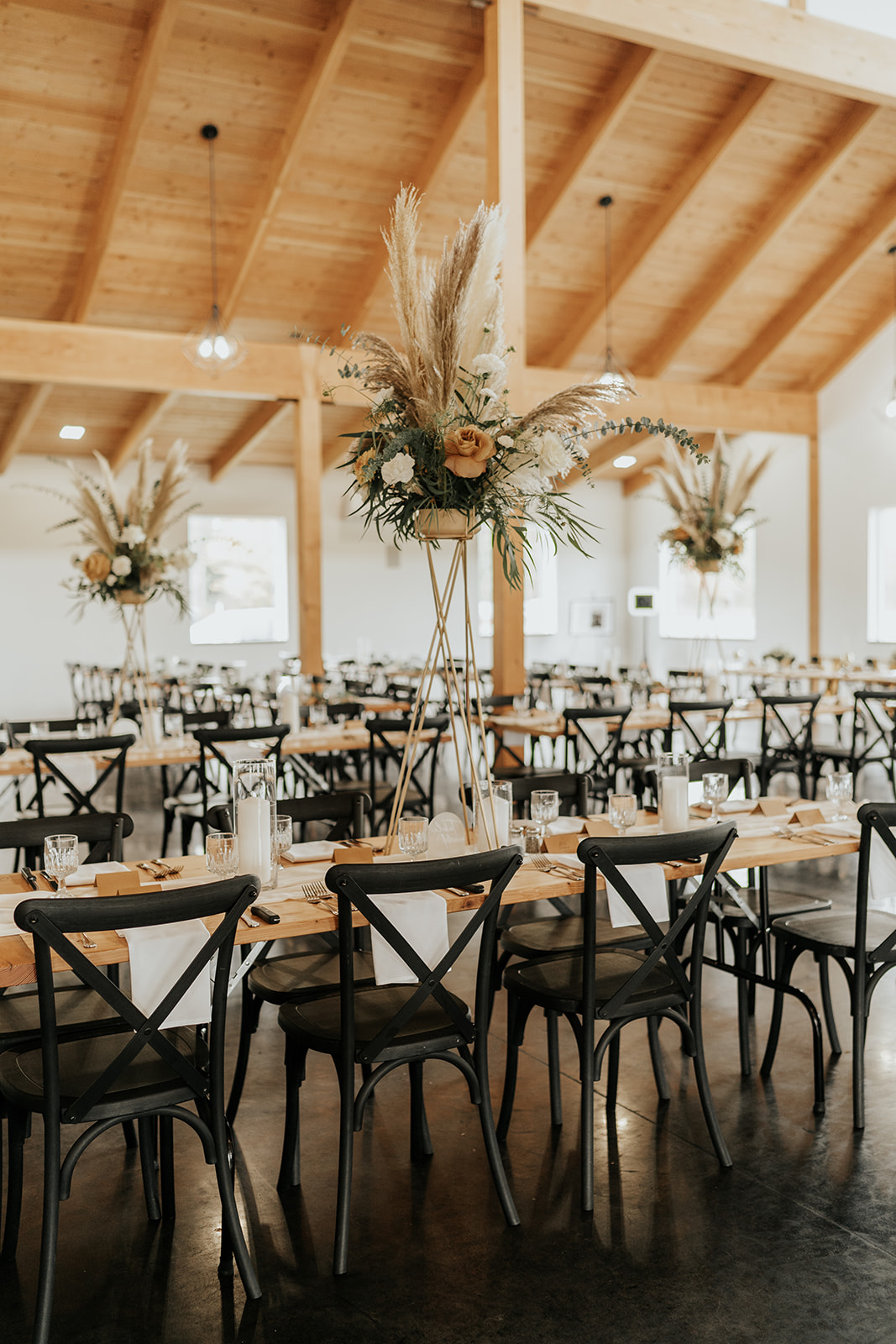 Elegant wedding reception hall with wooden tables, black chairs, and tall floral centerpieces under a wooden beam ceiling.