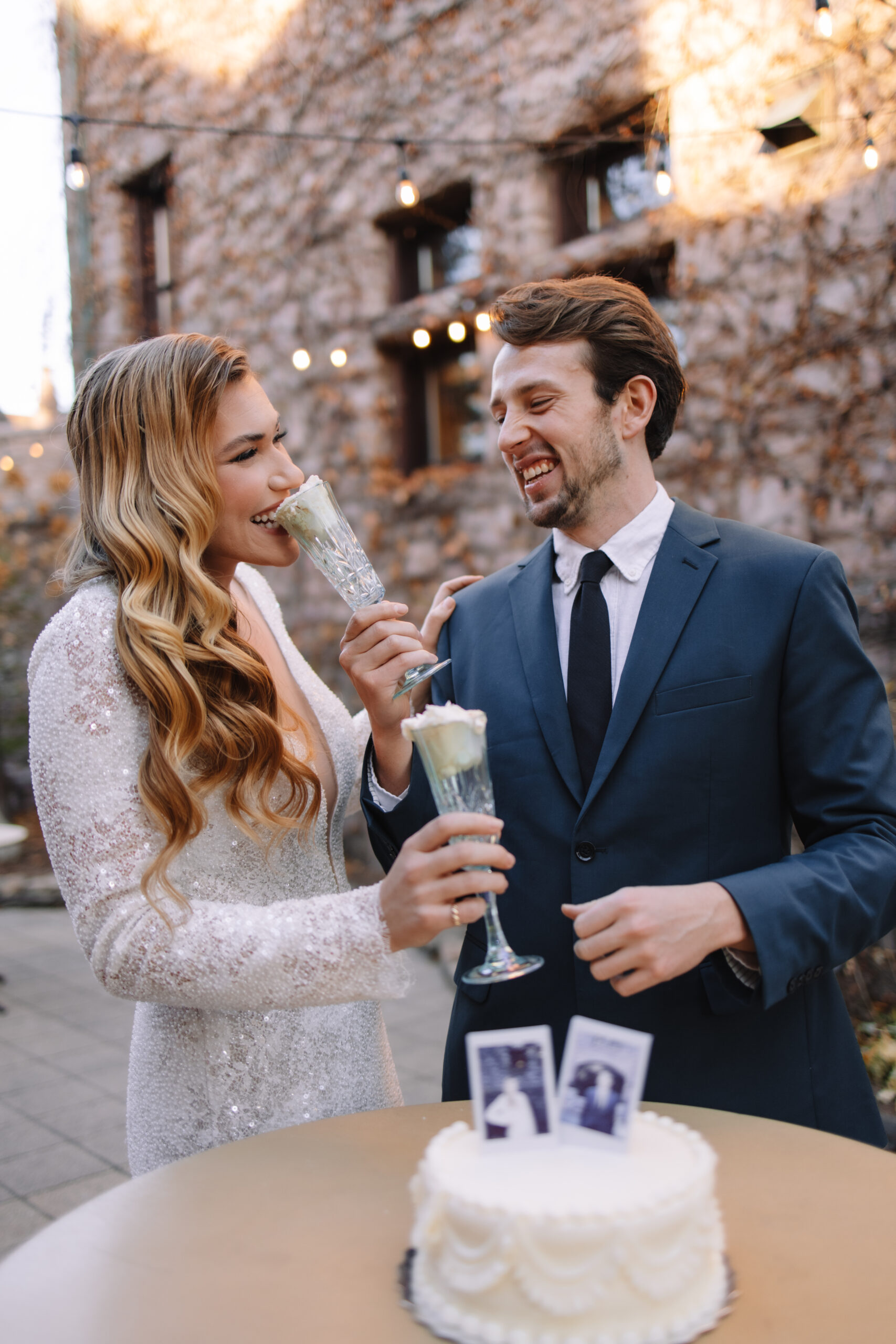 A bride and groom laugh and clink champagne glasses at an outdoor wedding reception, with a small cake and photos on the table in front of them. Her dress from a wedding dress shop in minneapolis 