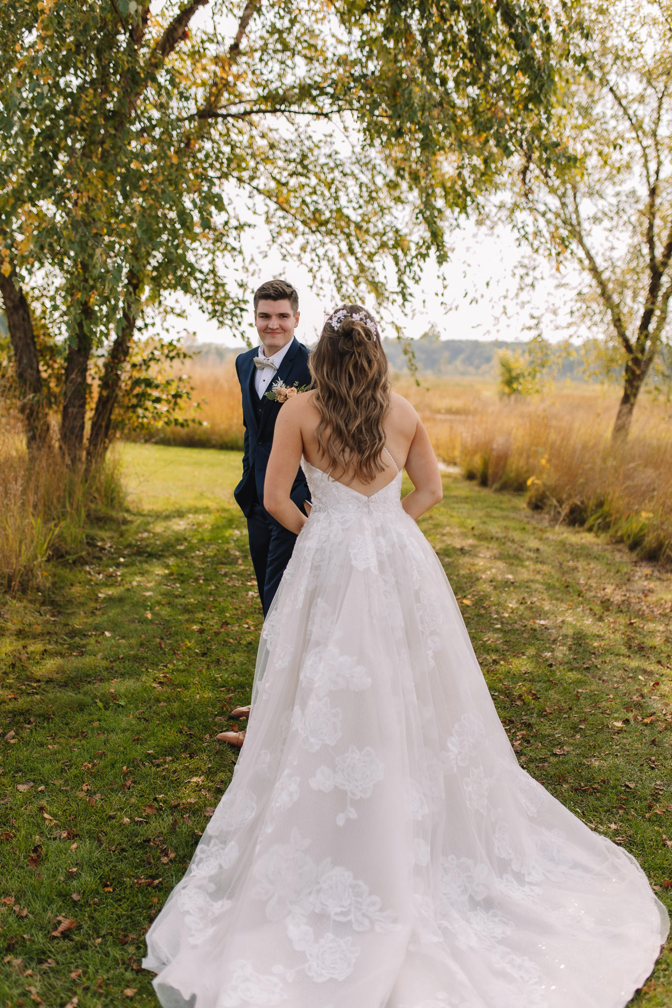 A bride in a white lace gown and a groom in a navy suit walking hand in hand in a grassy field, smiling.