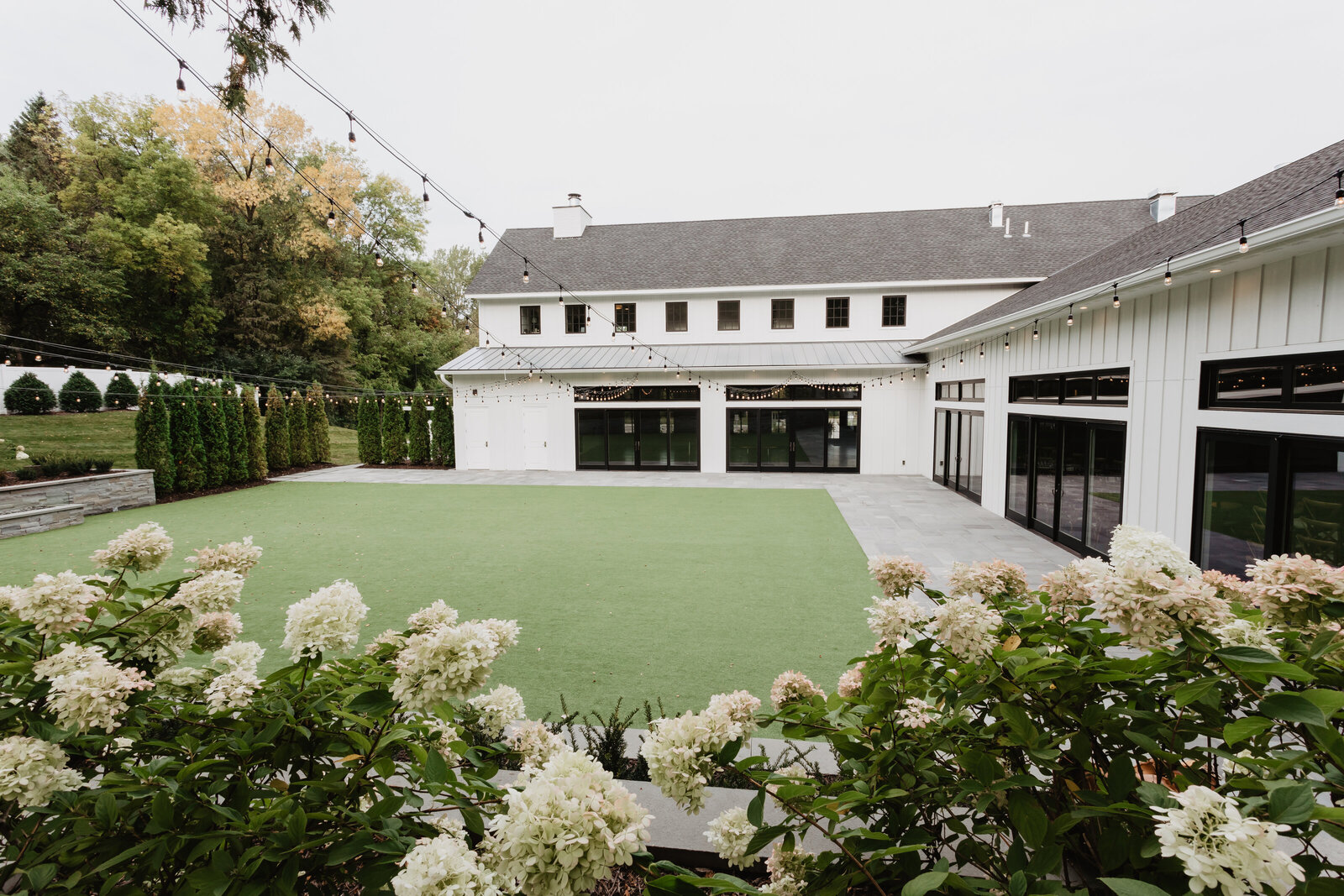A large modern wedding venue white building with black accents and a green synthetic lawn, surrounded by lush greenery and white flowers. This venue is called Hutton House