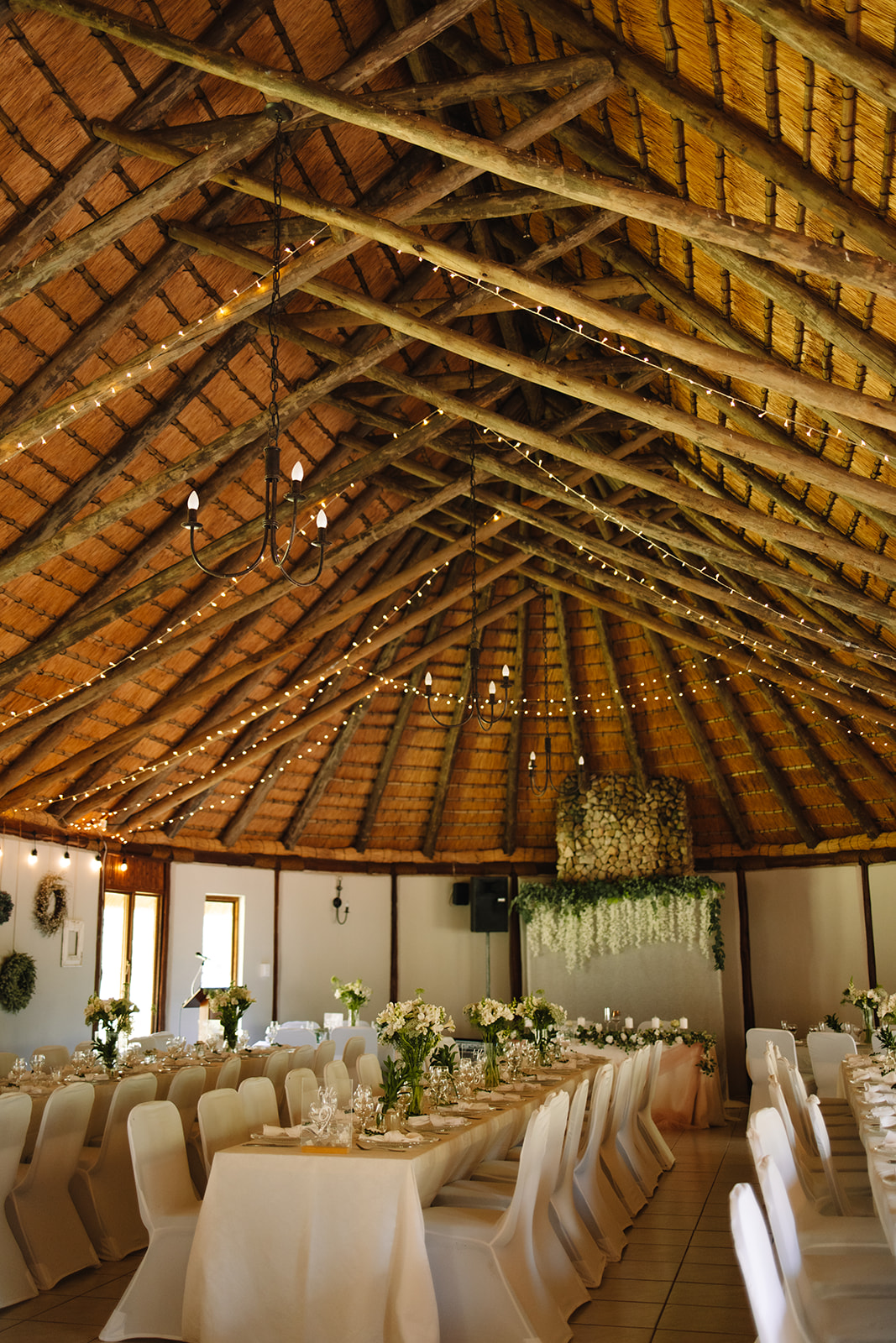Interior of a rustic event hall with high wooden beam ceiling, decorated with fairy lights and chandeliers, set up for a wedding reception with white draped tables at a wedding in Minnesota 