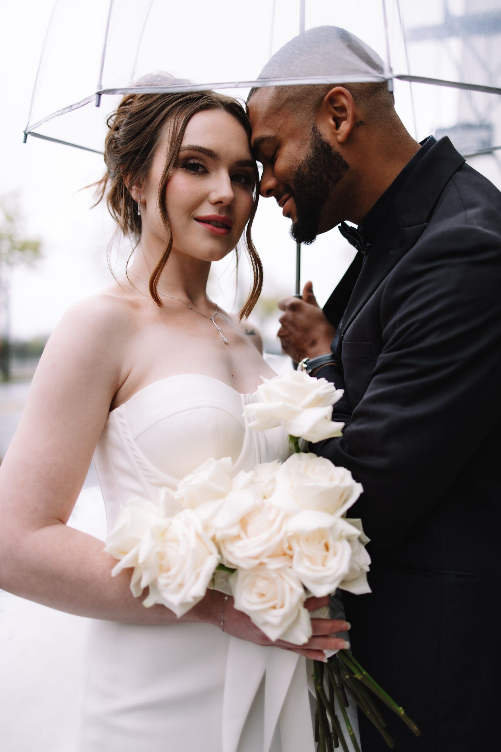 Bride and groom standing underneath a clear umbrella in the mist rain with a  simple white rose bouquet at the Machine Shop Wedding venue in Minneapolis