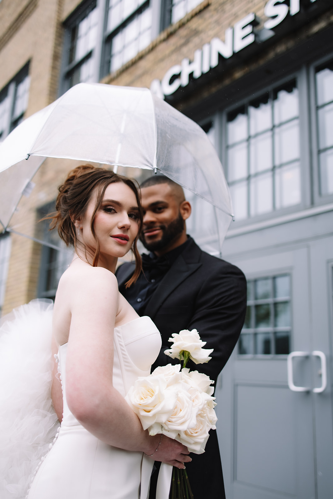 A bride in a white dress holding white roses and a groom in a black suit stand under a transparent umbrella on a rainy day in front of a brick building at the machine shop in minneapolis