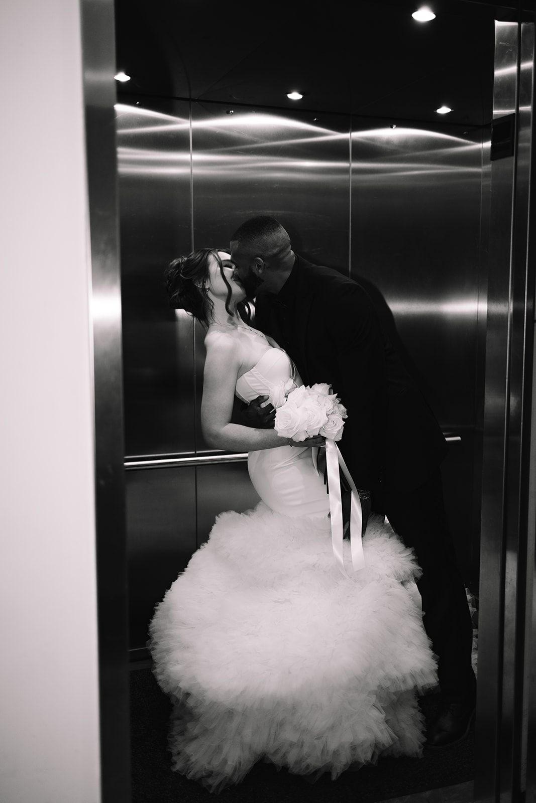 A bride and groom share a kiss in an elevator. The bride is wearing a white wedding dress and holding a bouquet of flowers, while the groom is in a black suit at the machine shop in minneapolis