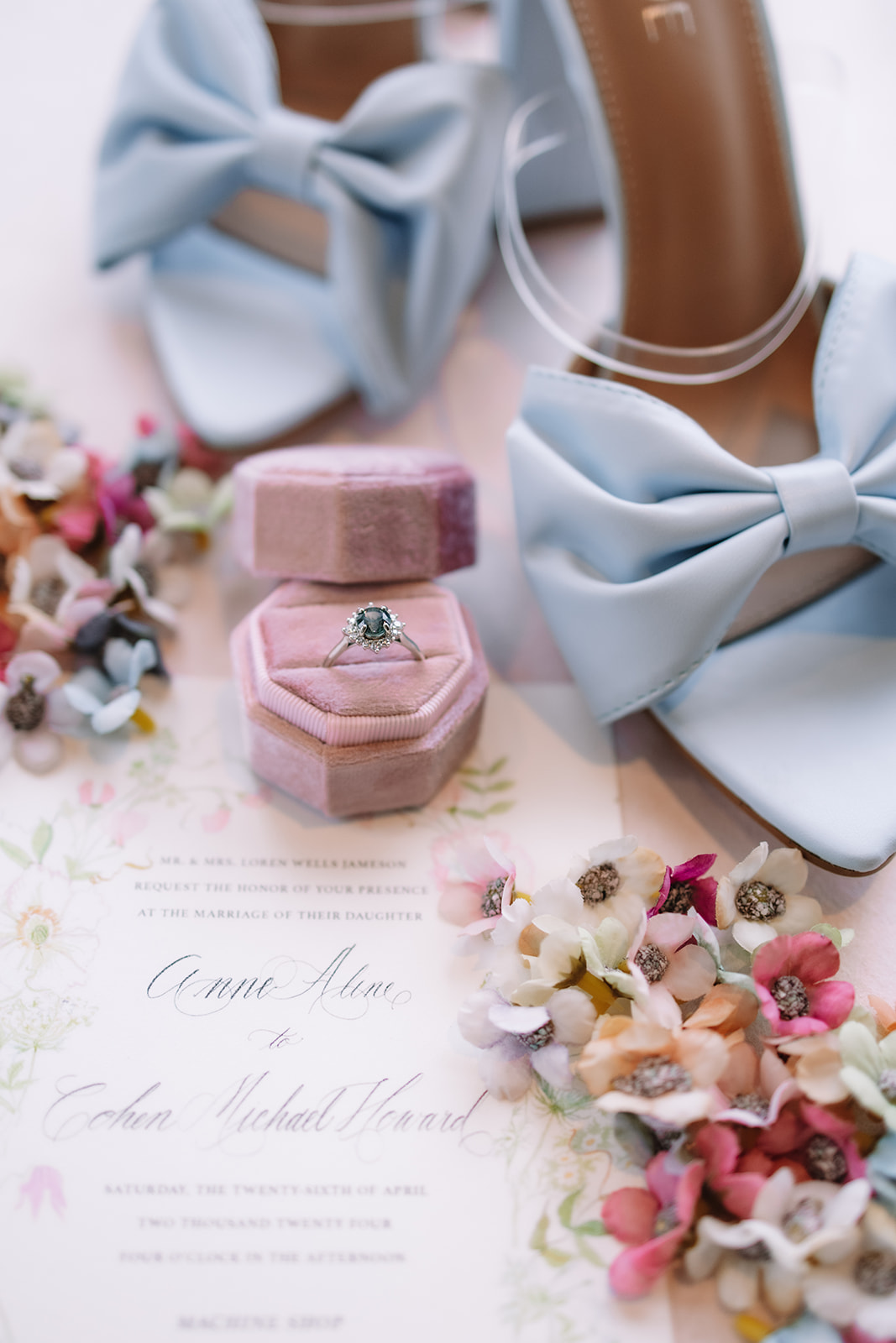 A colorful pastel flat lay wedding detail shot of baby blue wedding heels with bows, a blush ring holder with a ring in it and a wedding invitation surrounded by small pastel flowers