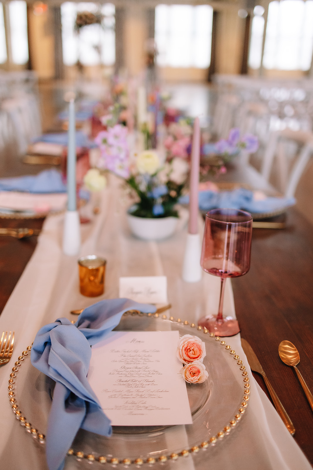 A table setting with a clear glass plate, a blue cloth napkin, a menu, a pink wine glass, and small pink roses. The table features pink and purple flower arrangements and pink and blue candles.