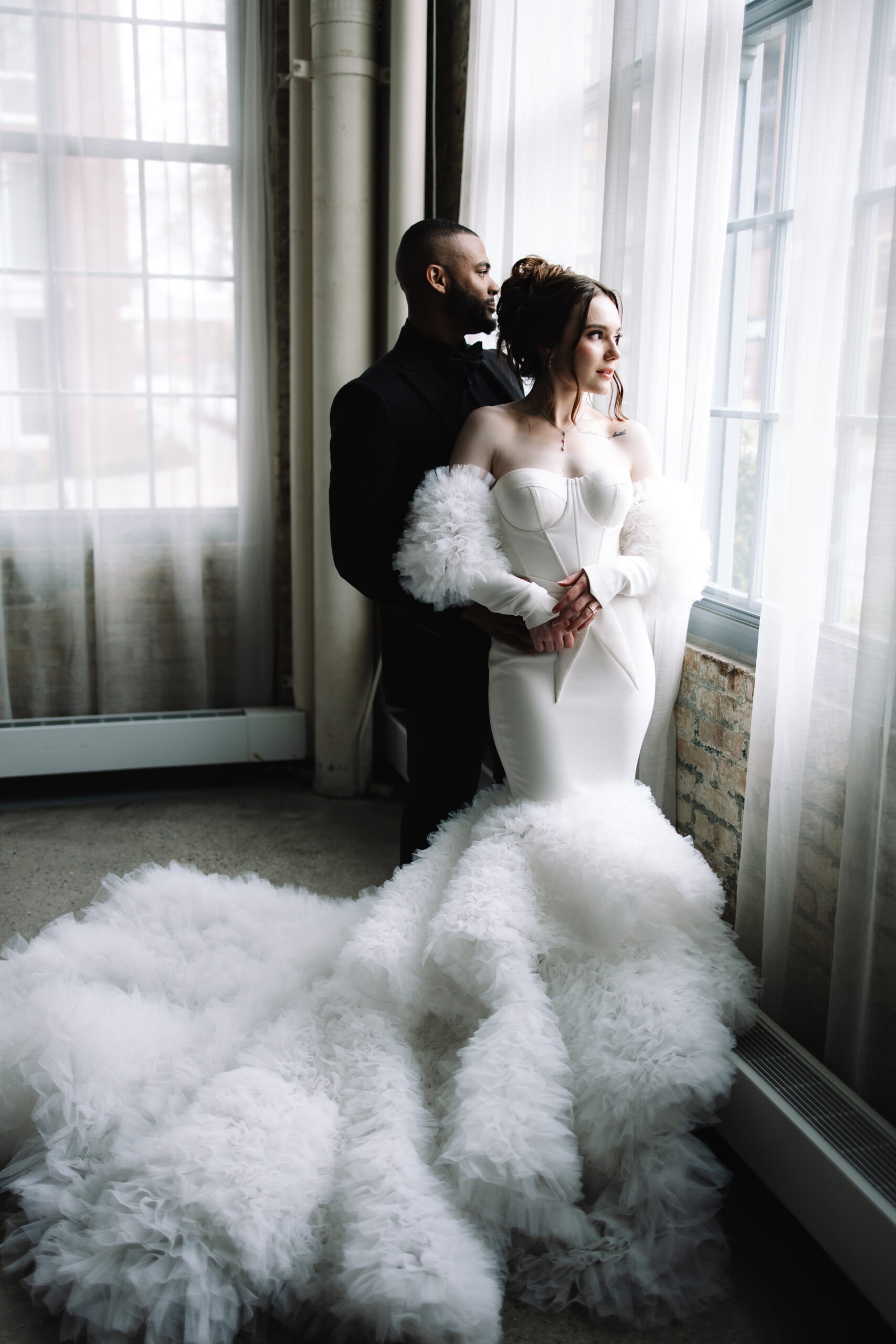 A Groom in a black suit holding his bride from behind as they stare out the large window. The bride is wearing a voluptuous mermaid wedding dress.