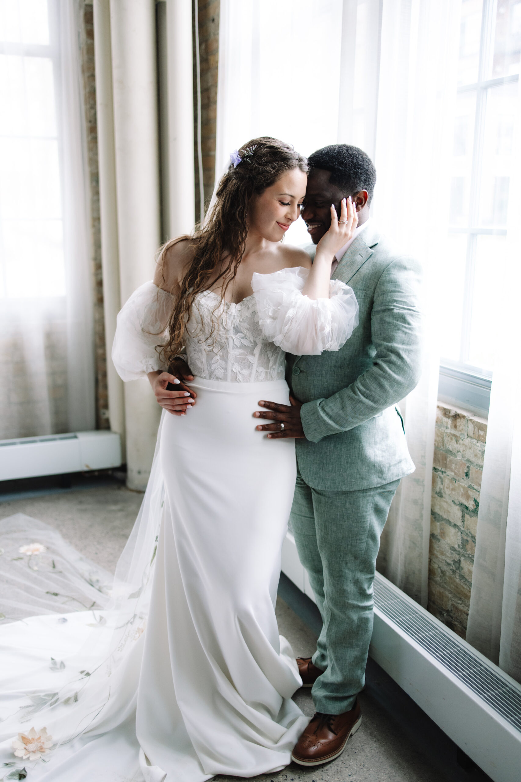 A bride and groom embrace lovingly near a window, the bride in a white dress and the groom in a gray suit.