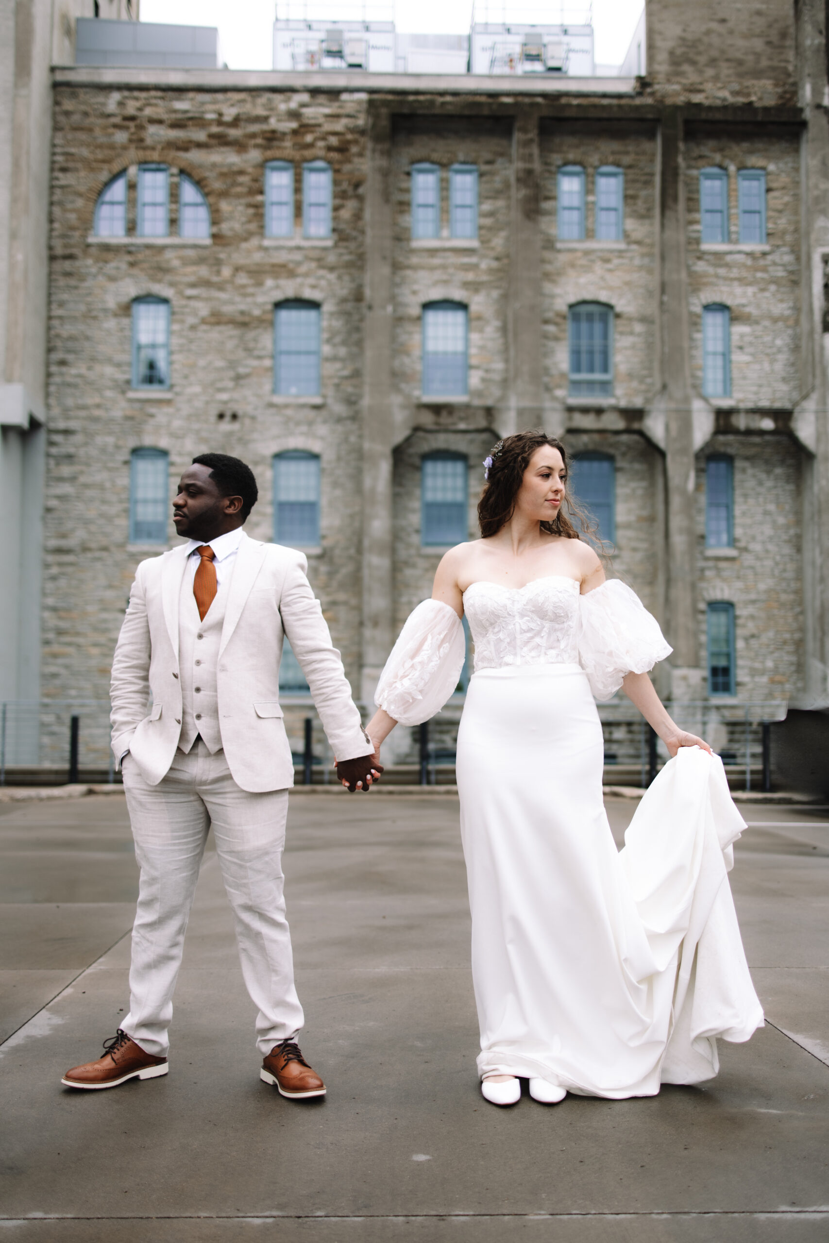 A bride in a white dress and a groom in a white suit hold hands, standing back to back in front of a stone building.