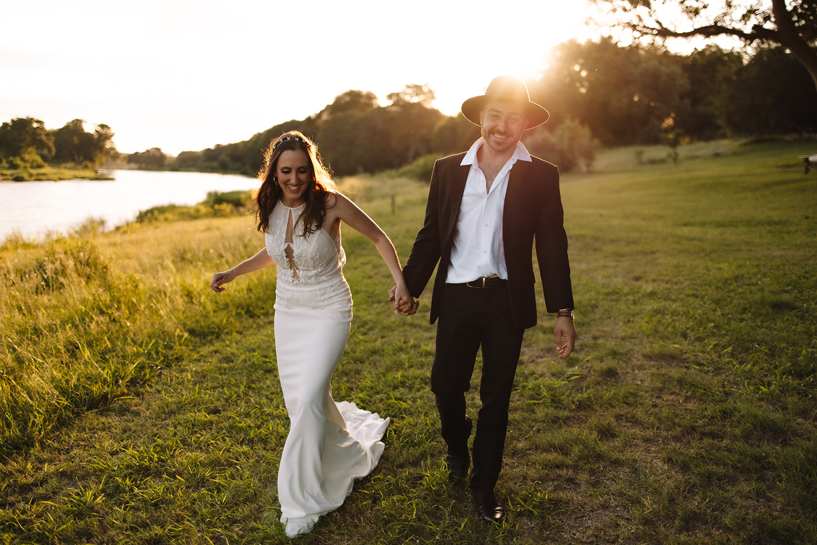 A couple dressed in wedding attire, with the groom in a hat, holding hands and walking on a grassy field by a river at sunset.