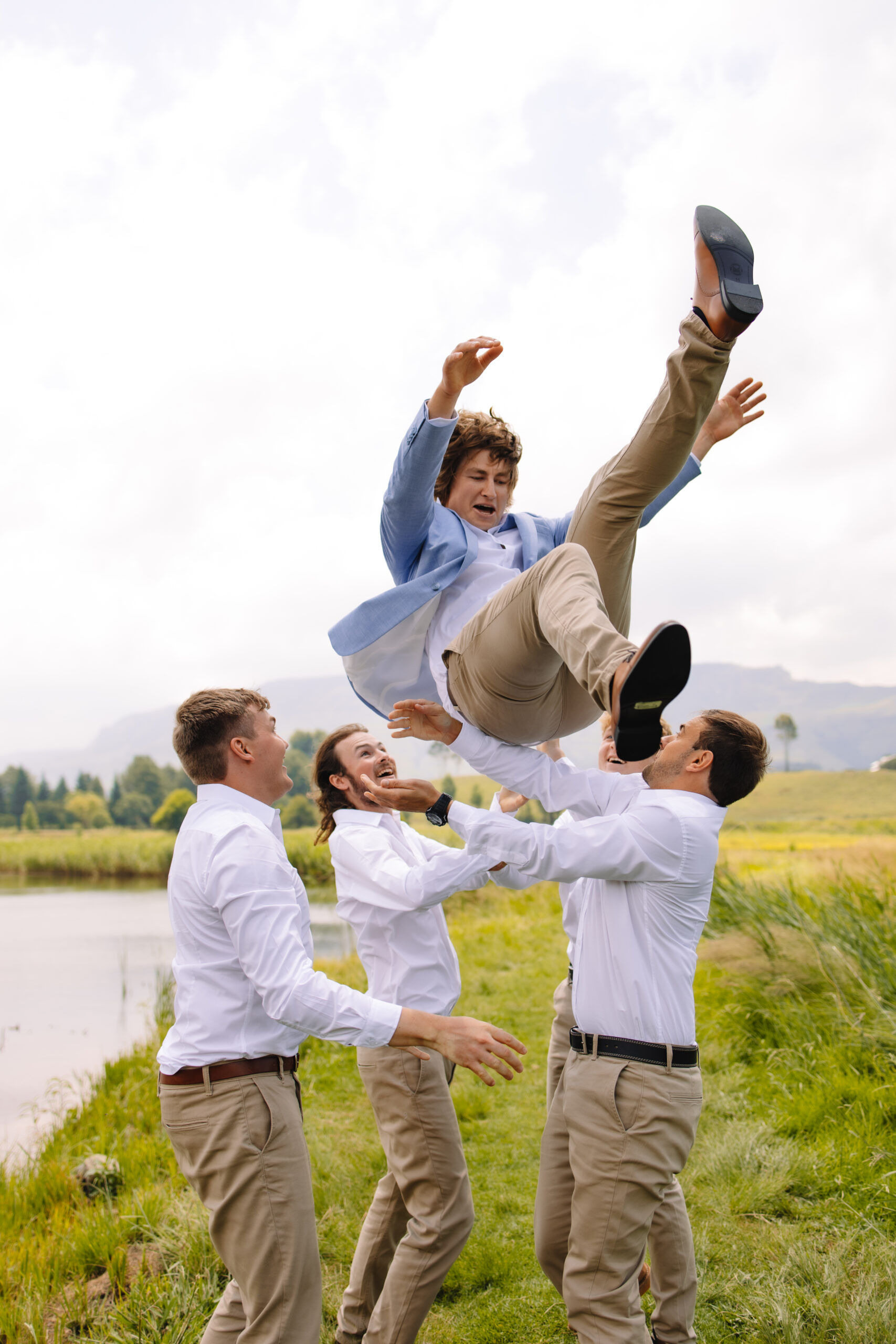 Groomsmen in white button up shirts and khaki pants throwing the groom in the air next to a lake.