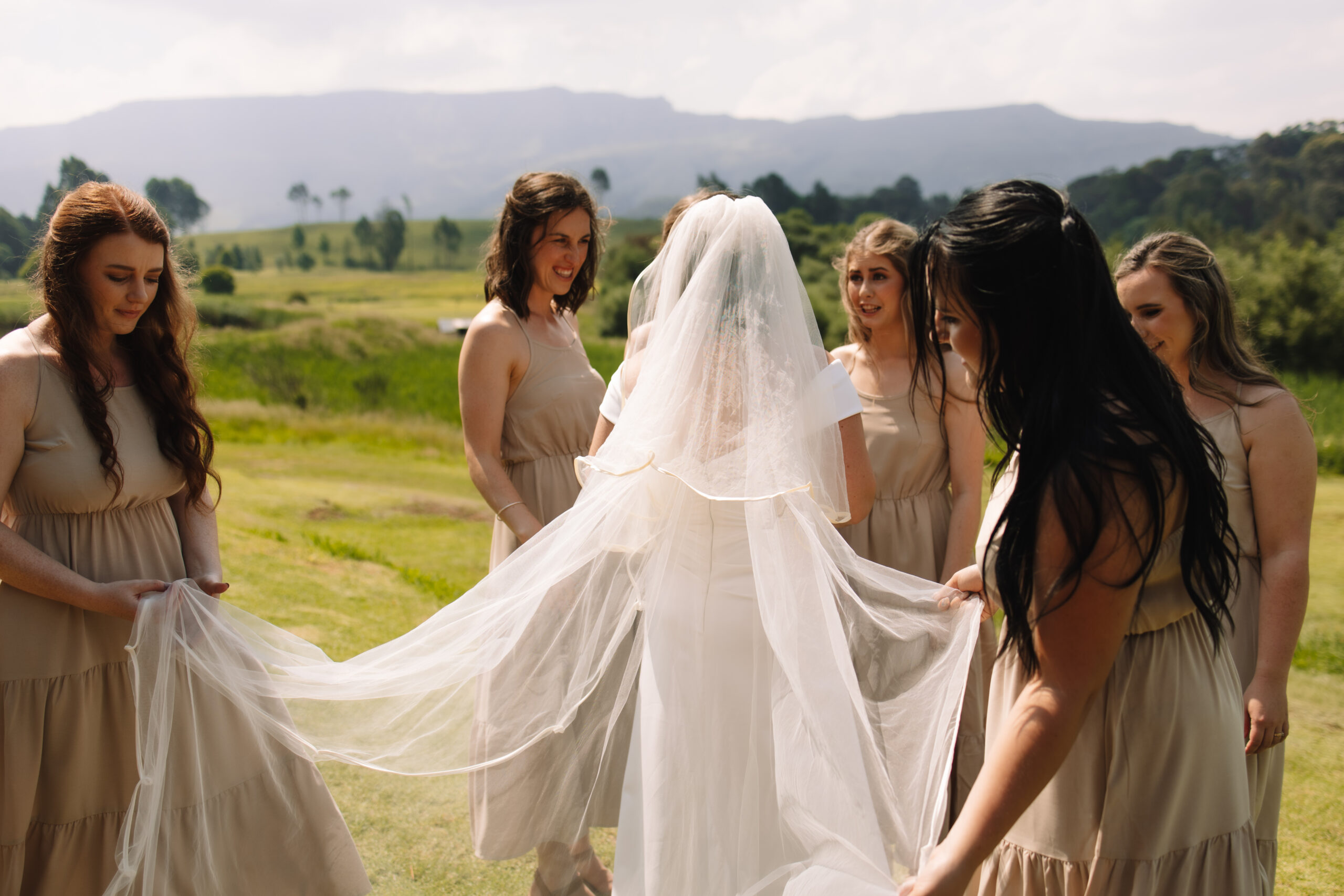 A bride with a long veil, standing outside surrounded by her bridesmaids holding her veil and admiring how beautiful she looks