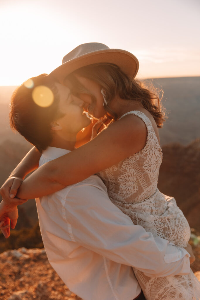 Groom holding his bride and going in for a delicate kiss as the sun sets behind them. The bride is wearing a hat and a lace wedding dress