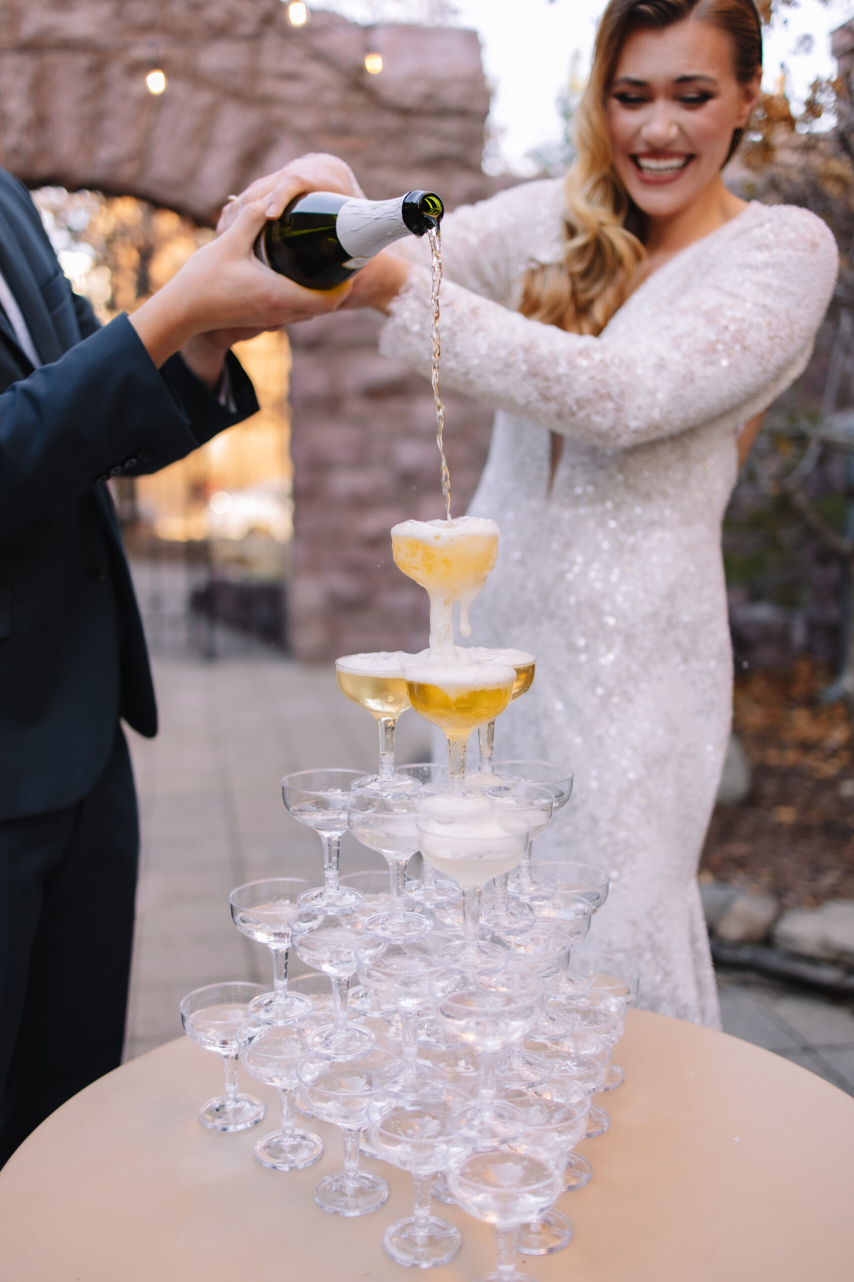 Bride in a sparkly long sleeve wedding dress pouring champagne into the top glass of the champagne tower