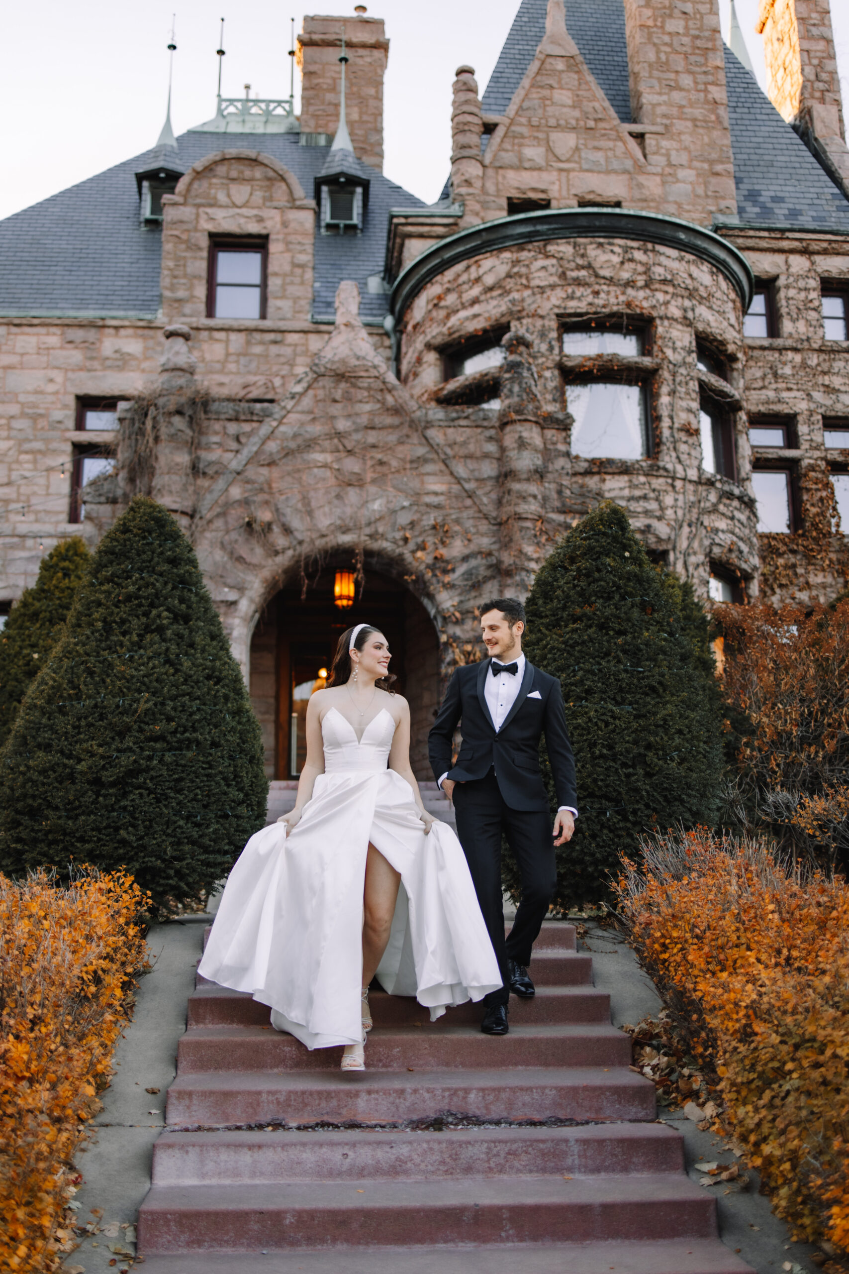 A couple in formal wedding attire walking down the steps of an ornate castle-like building called the Van Dusen Mansion in Minneapolis