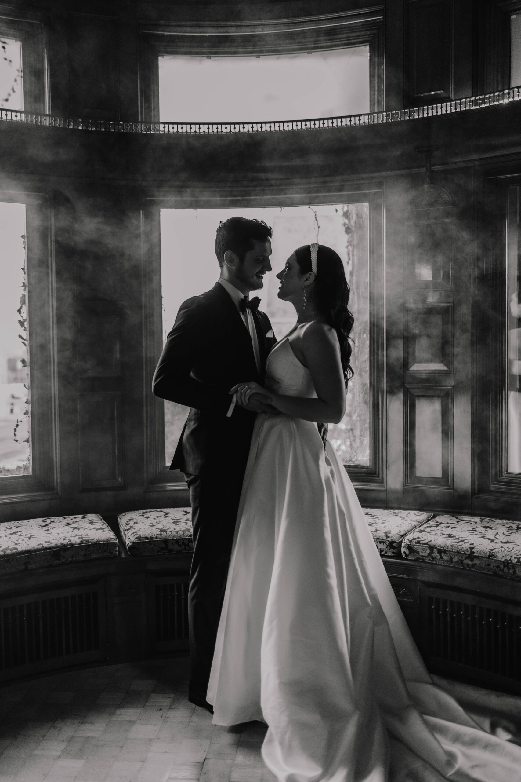 A couple in formal attire stand closely in an elegant wooden library. The woman wears a white gown and the man a black suit. They gaze at each other and appear to be in a romantic moment at the van dusen mansion