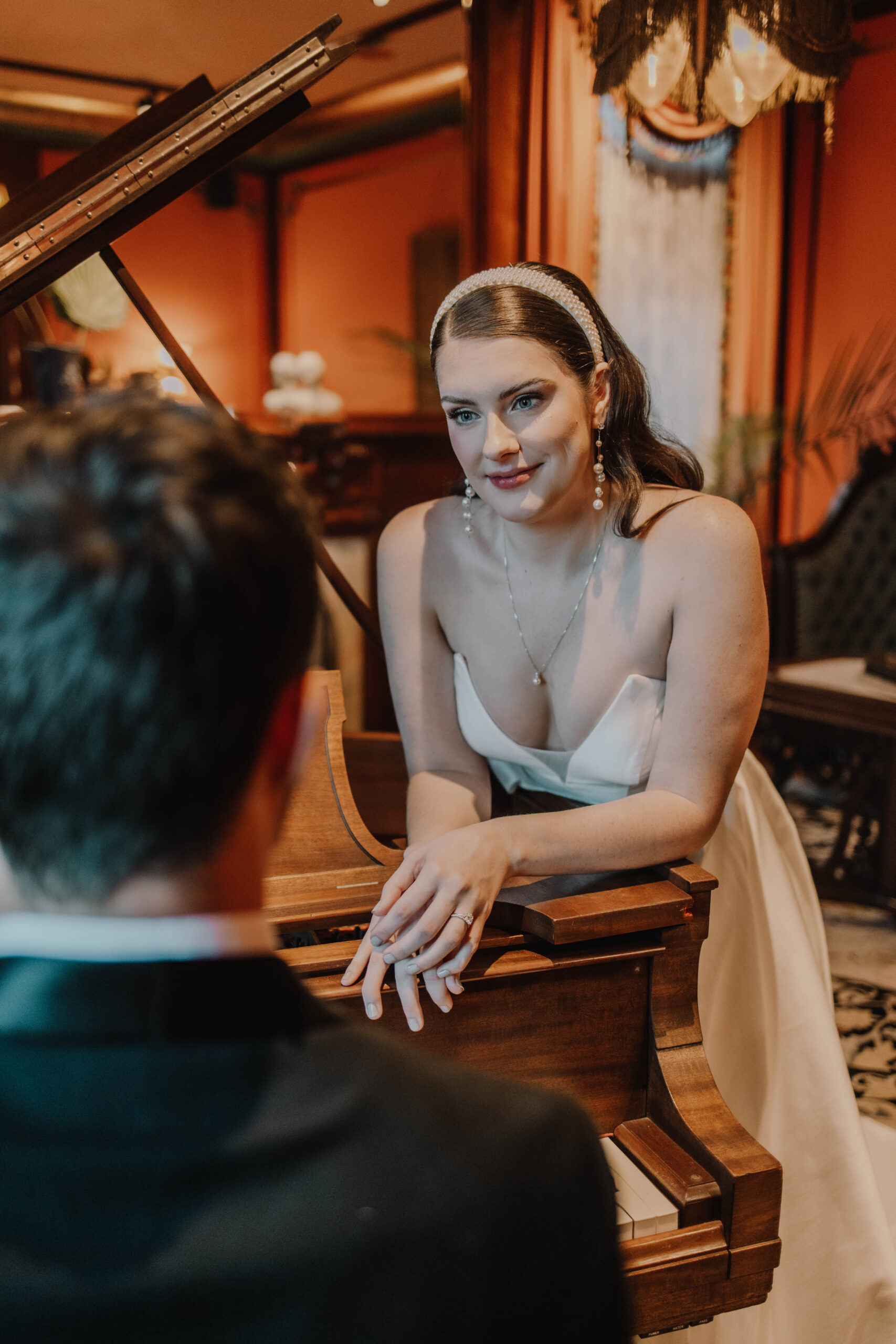 A bride leaning on a piano inside a mansion while her groom is playing her a song. She stares at him lovingly. 