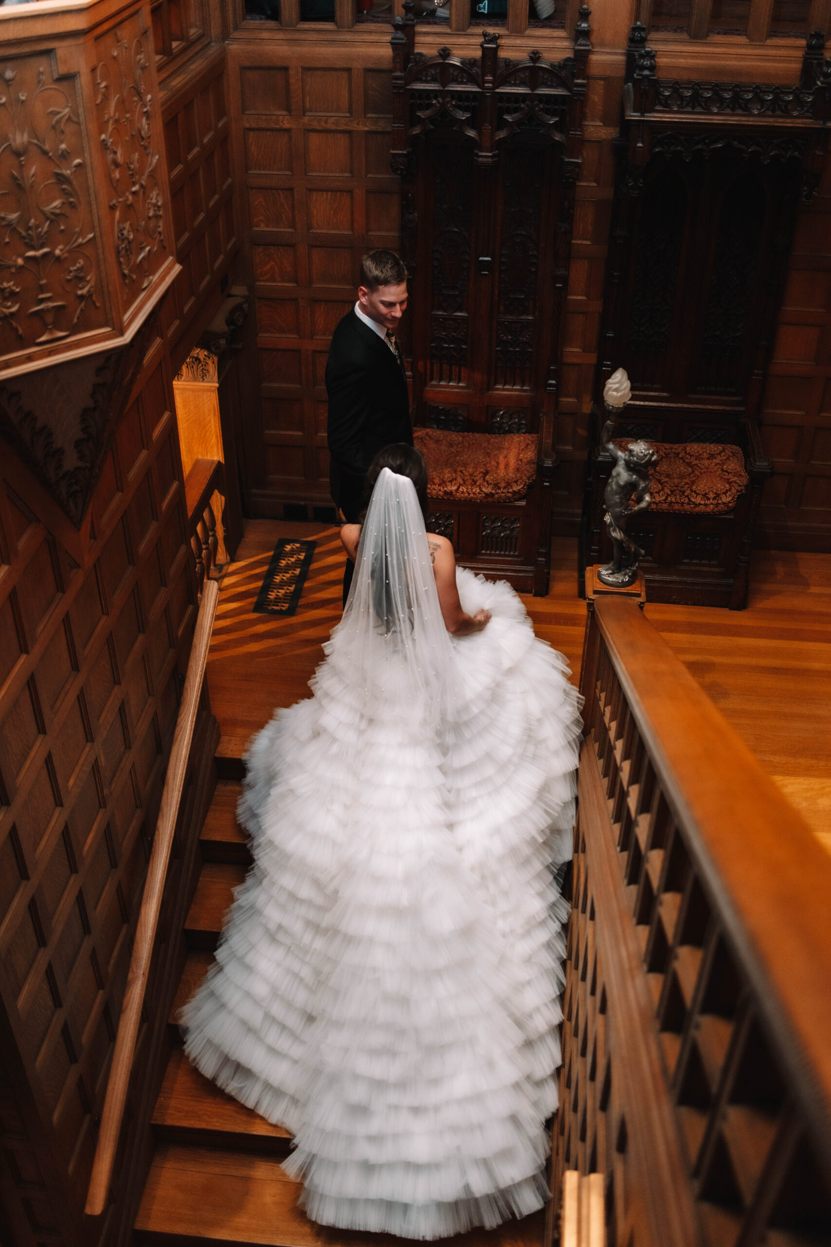A couple descended a wooden staircase; the woman wore a voluminous white wedding gown with a long train, and the man is in a formal suit. The surroundings featured intricate wood paneling and carvings.