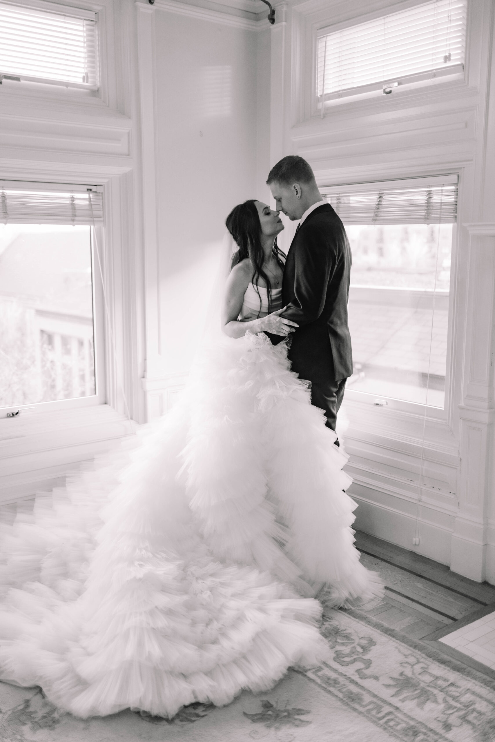 A bride in a white gown and a groom in a black suit share an intimate moment by a window in a softly lit room.